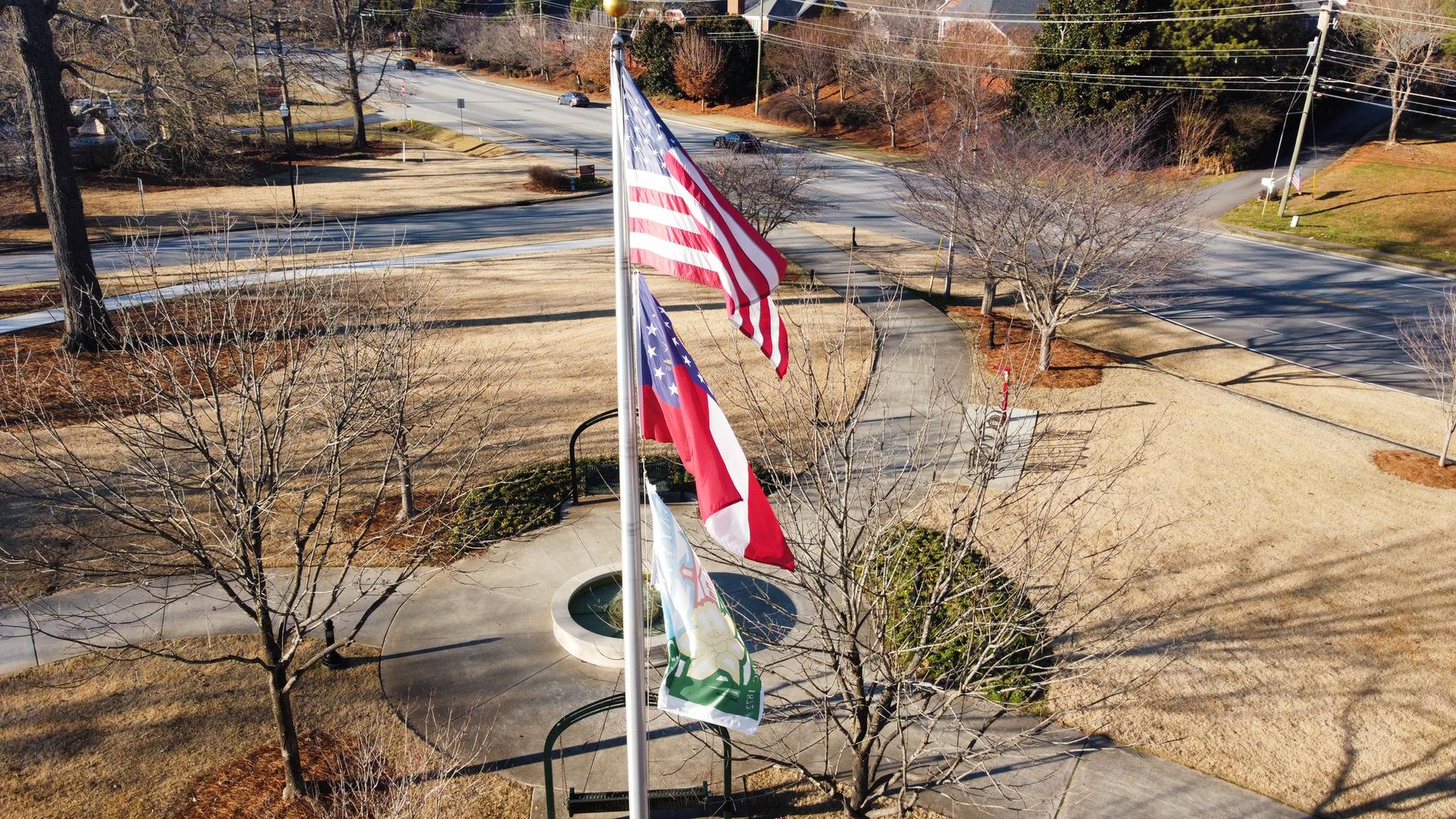 Three flags fly on poles in a sunny park with bare trees.