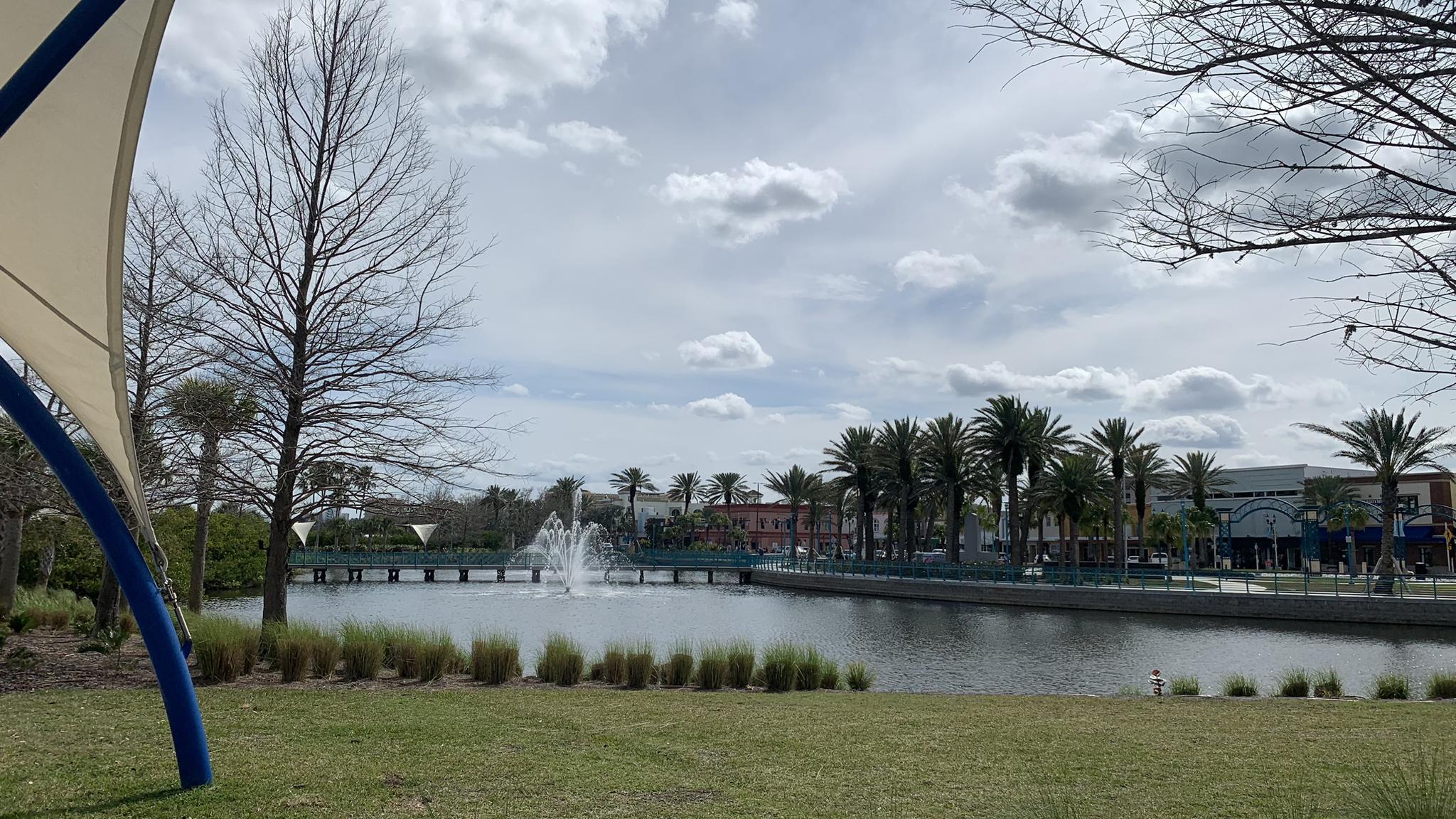 Pond with a fountain, palm trees, and buildings under a cloudy sky.