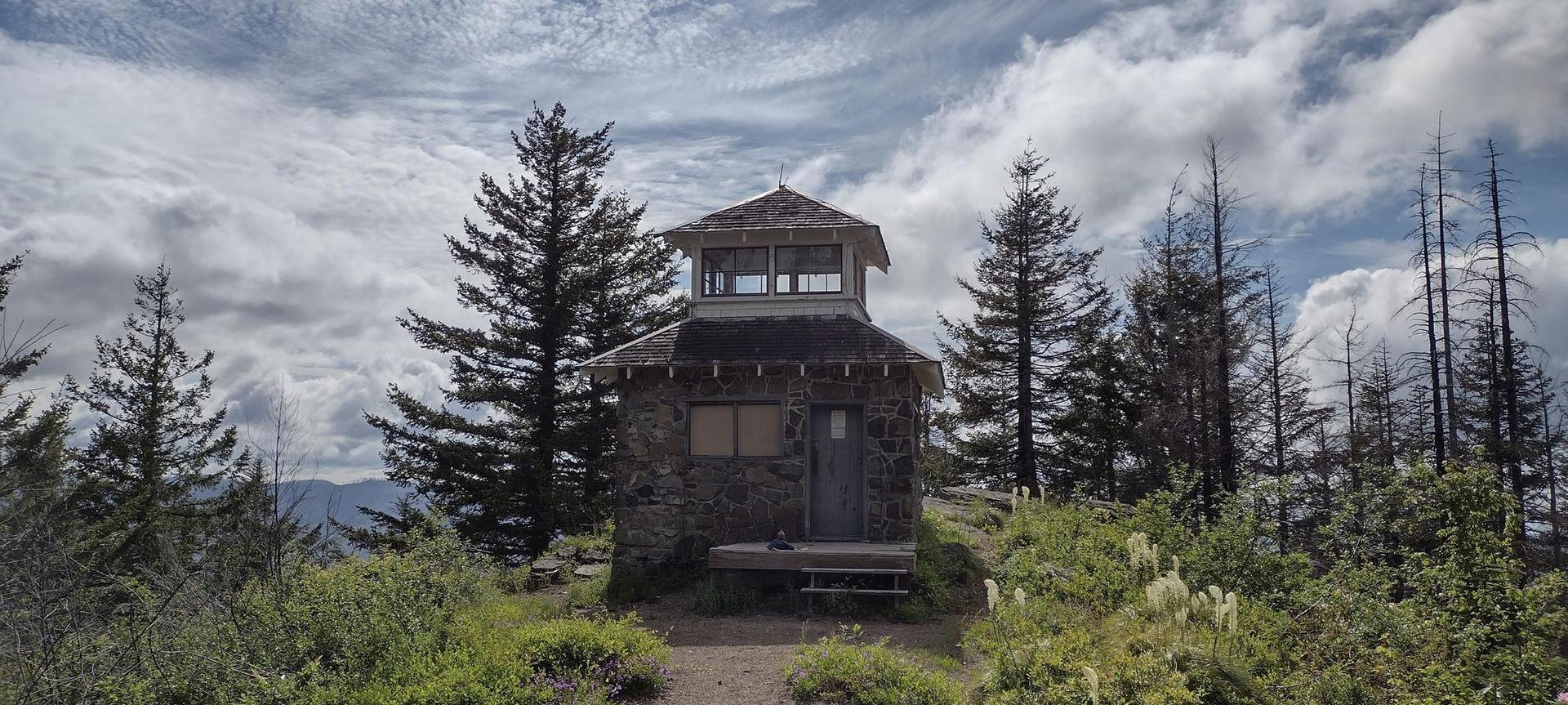 Stone lookout tower on a hilltop surrounded by tall trees, cloudy sky.