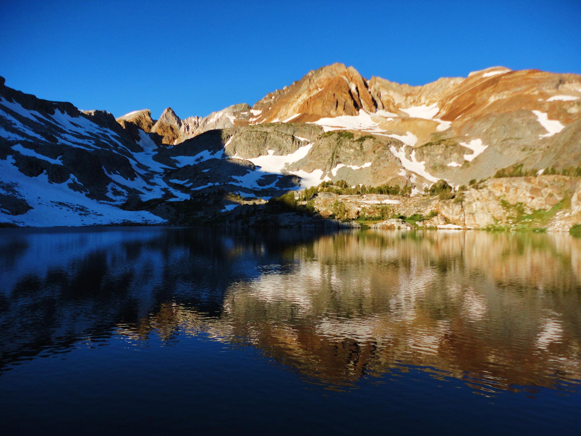 Dark lake reflecting rugged, snow-capped mountains bathed in warm, golden light.