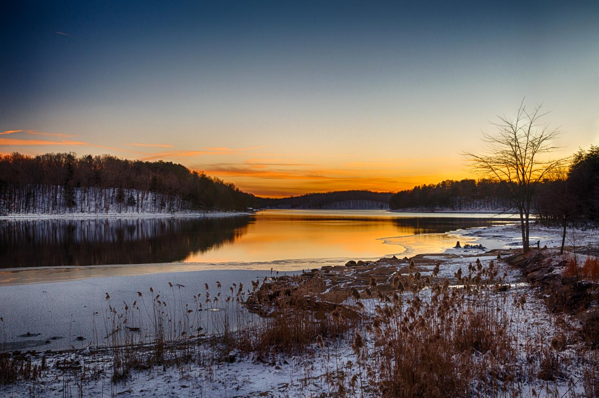 Winter sunset over a lake with snow-covered banks and trees reflecting in the water.