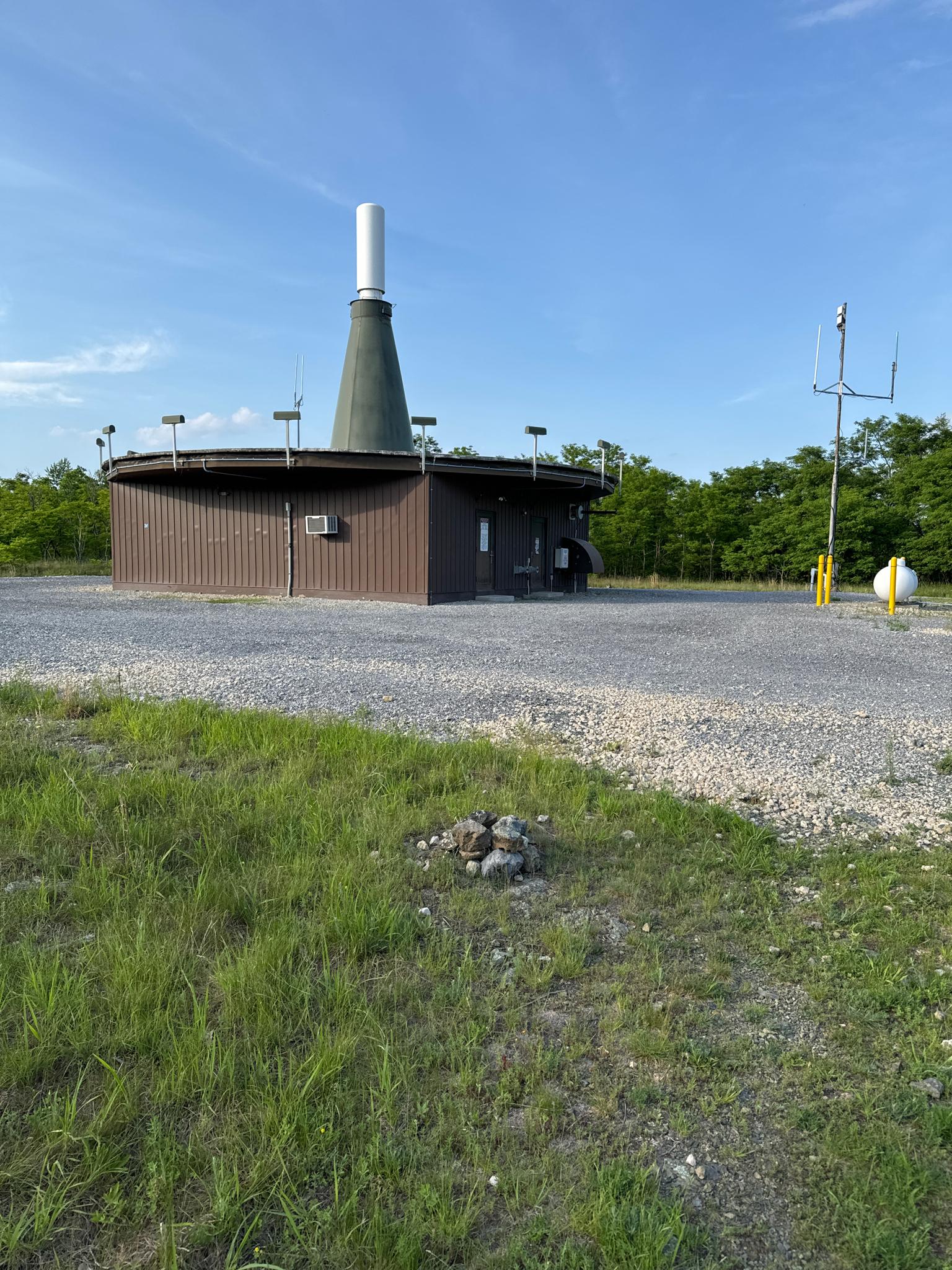Brown circular building with a tall white stack, surrounded by gravel and grass.