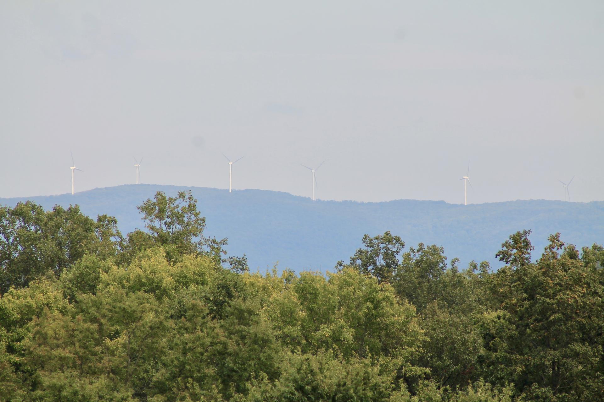 Distant wind turbines on a mountain ridge above green trees under a hazy sky.