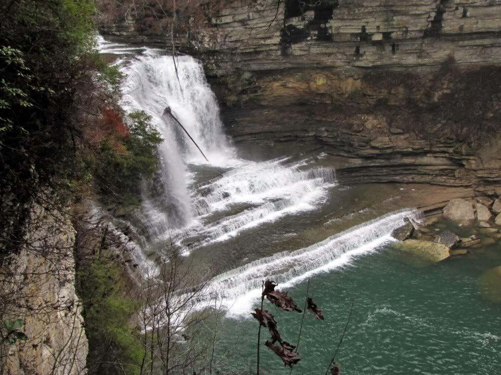A powerful waterfall cascades over rocky cliffs into a turquoise river under natural light.