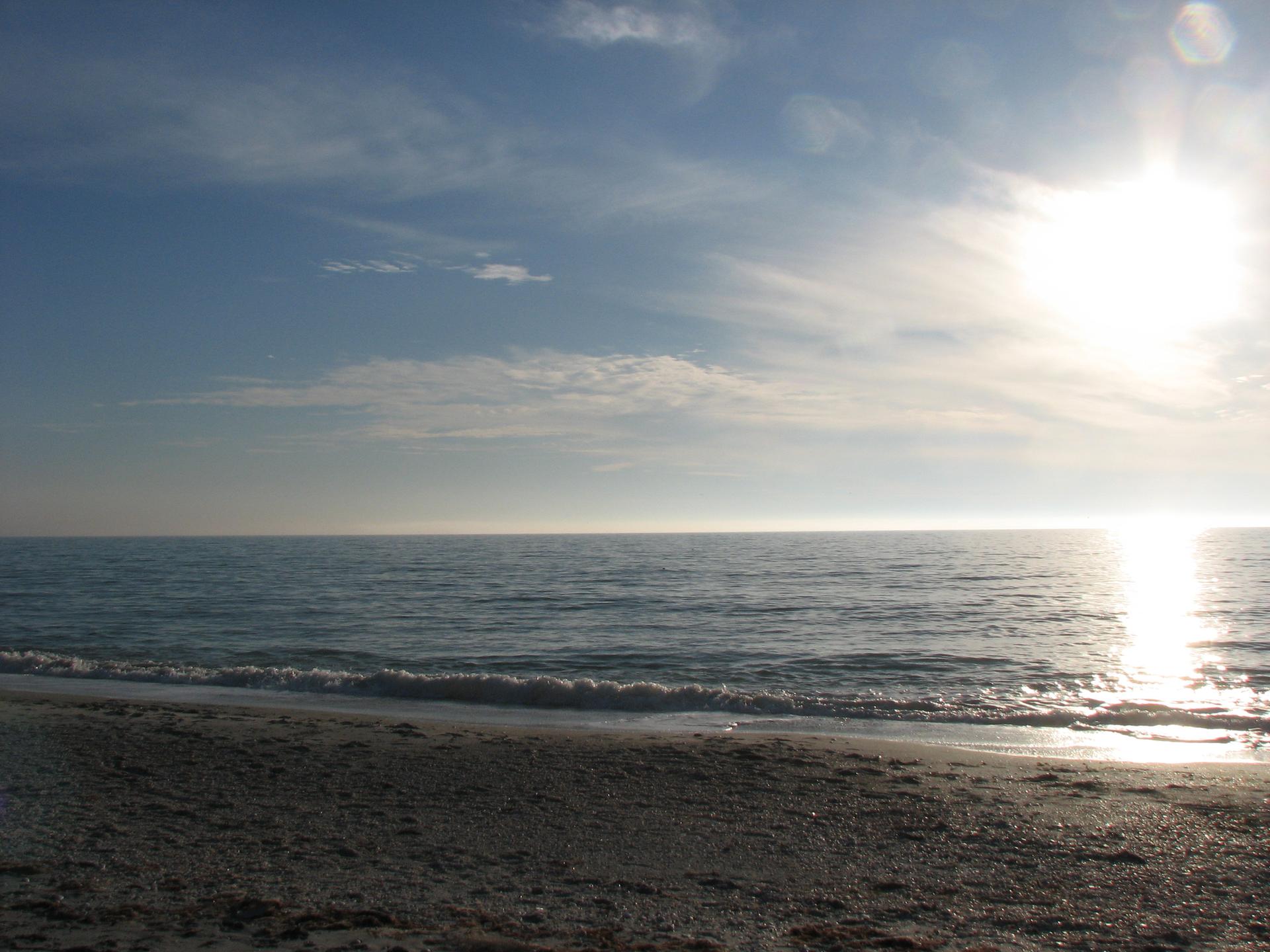 Bright sun reflecting on calm ocean water from a sandy beach.