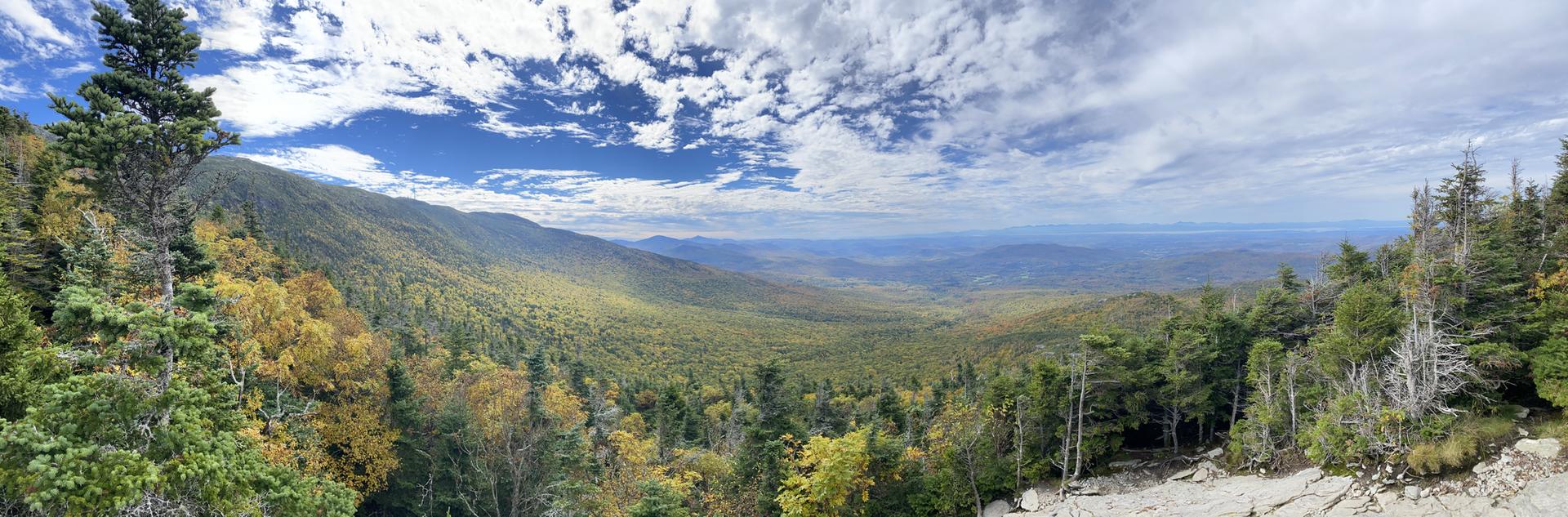 Expansive mountain vista with green and autumn trees under a bright, cloudy sky.