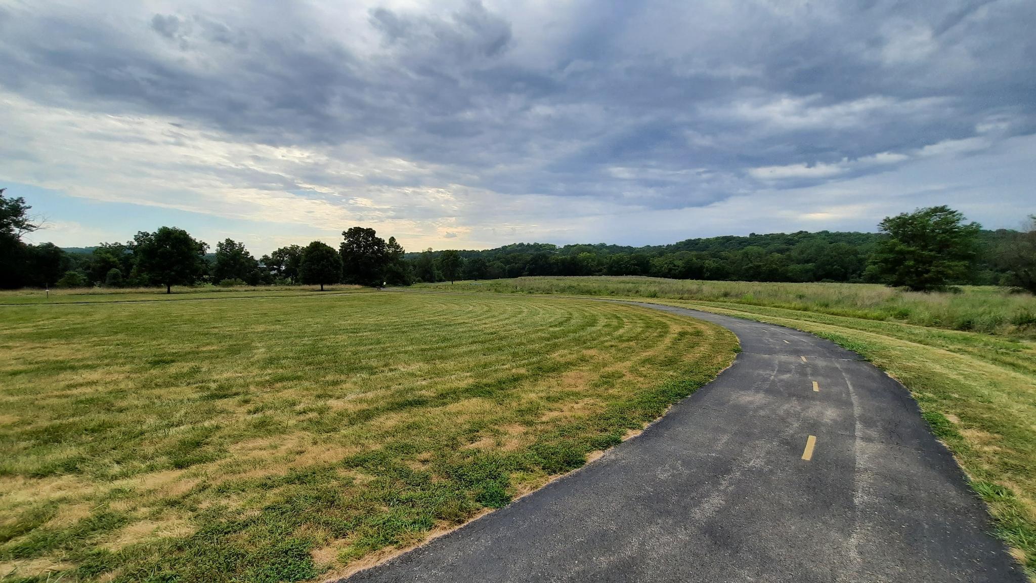 A paved path curves through a wide grassy field under a cloudy sky.