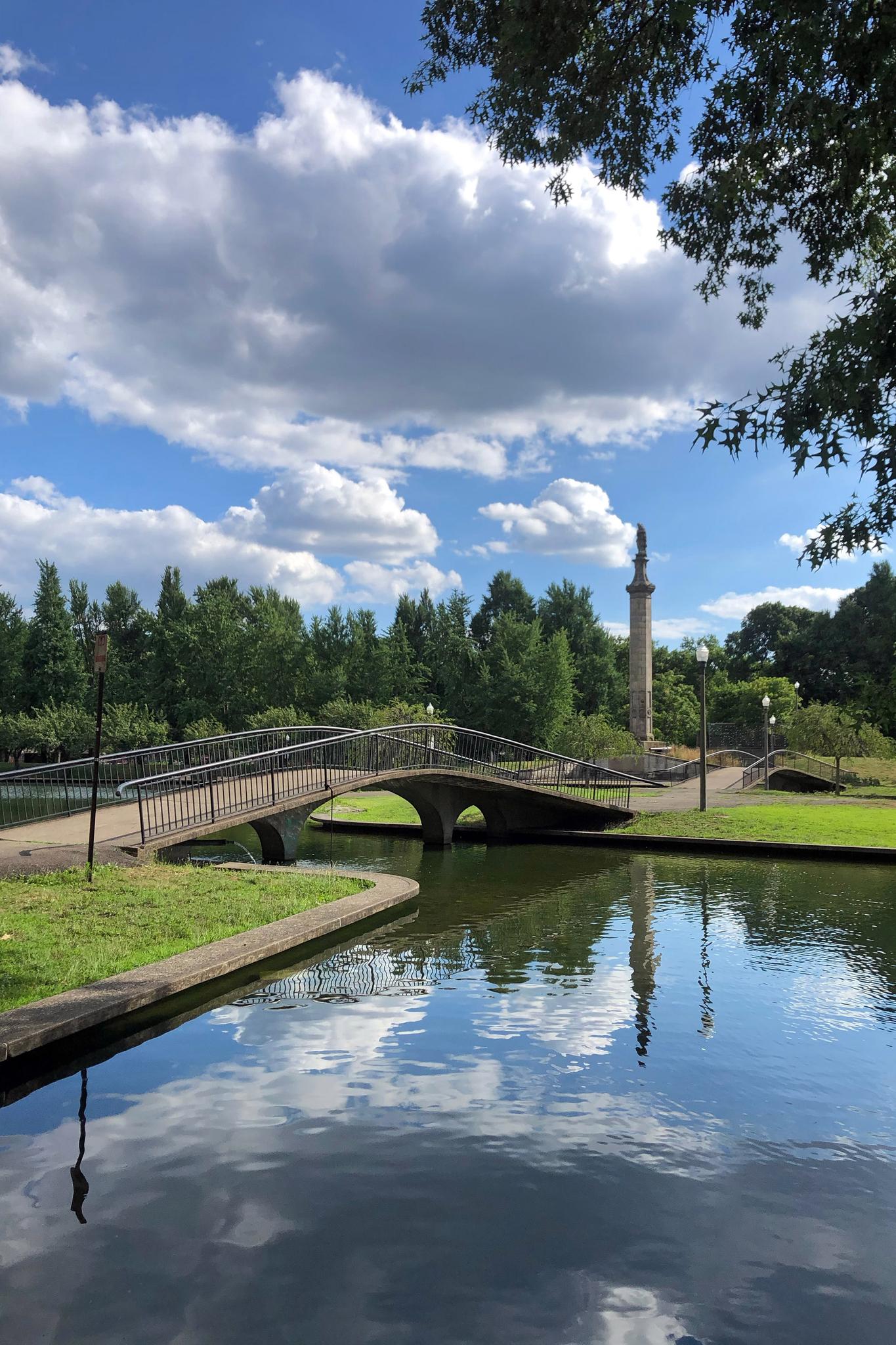 Arched bridge over a lake with a distant monument under a bright, cloudy sky.