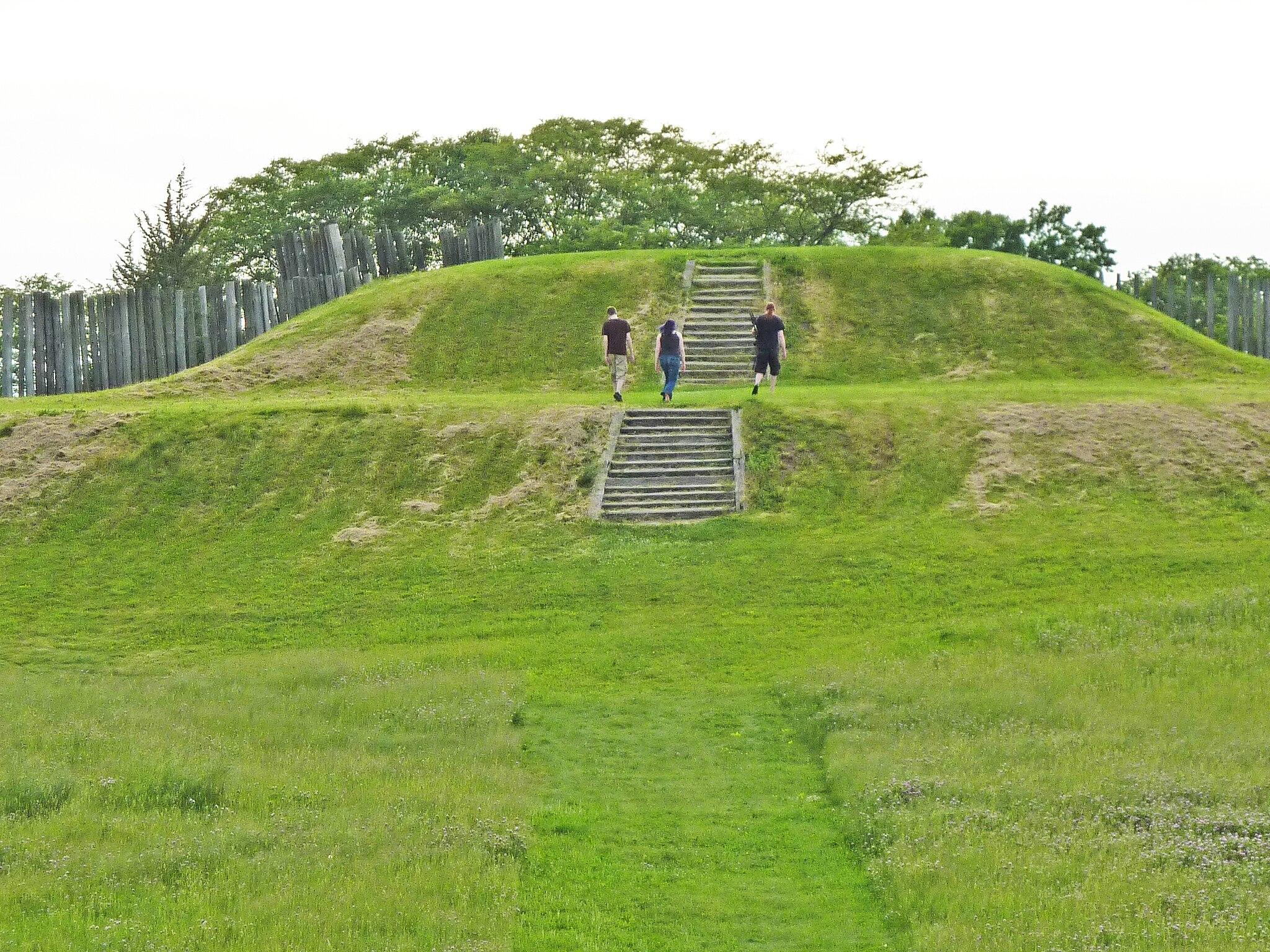 Three people ascend stone stairs on a large, sunny green grassy mound.