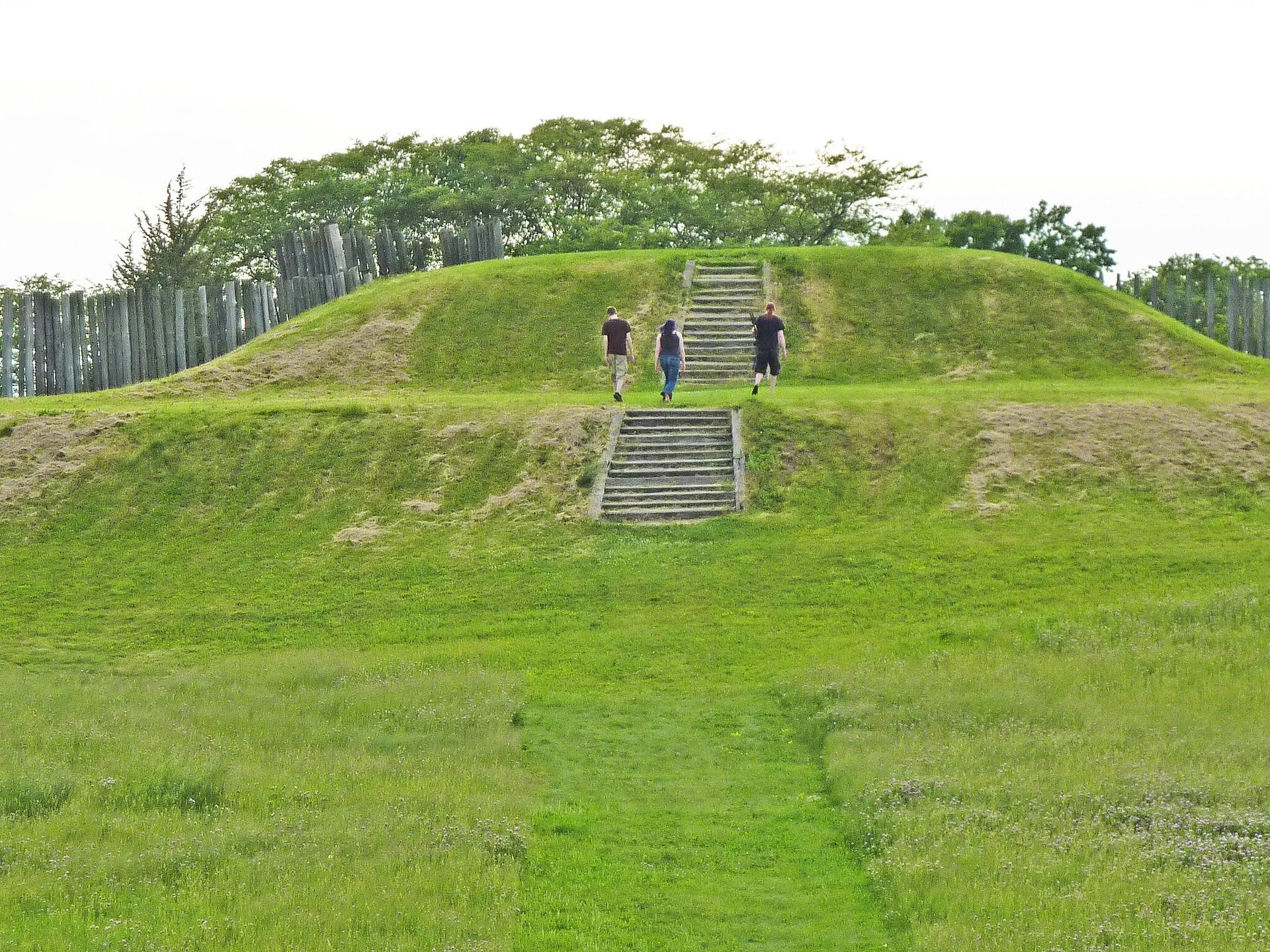 Three people ascend stone stairs on a large, sunny green grassy mound.