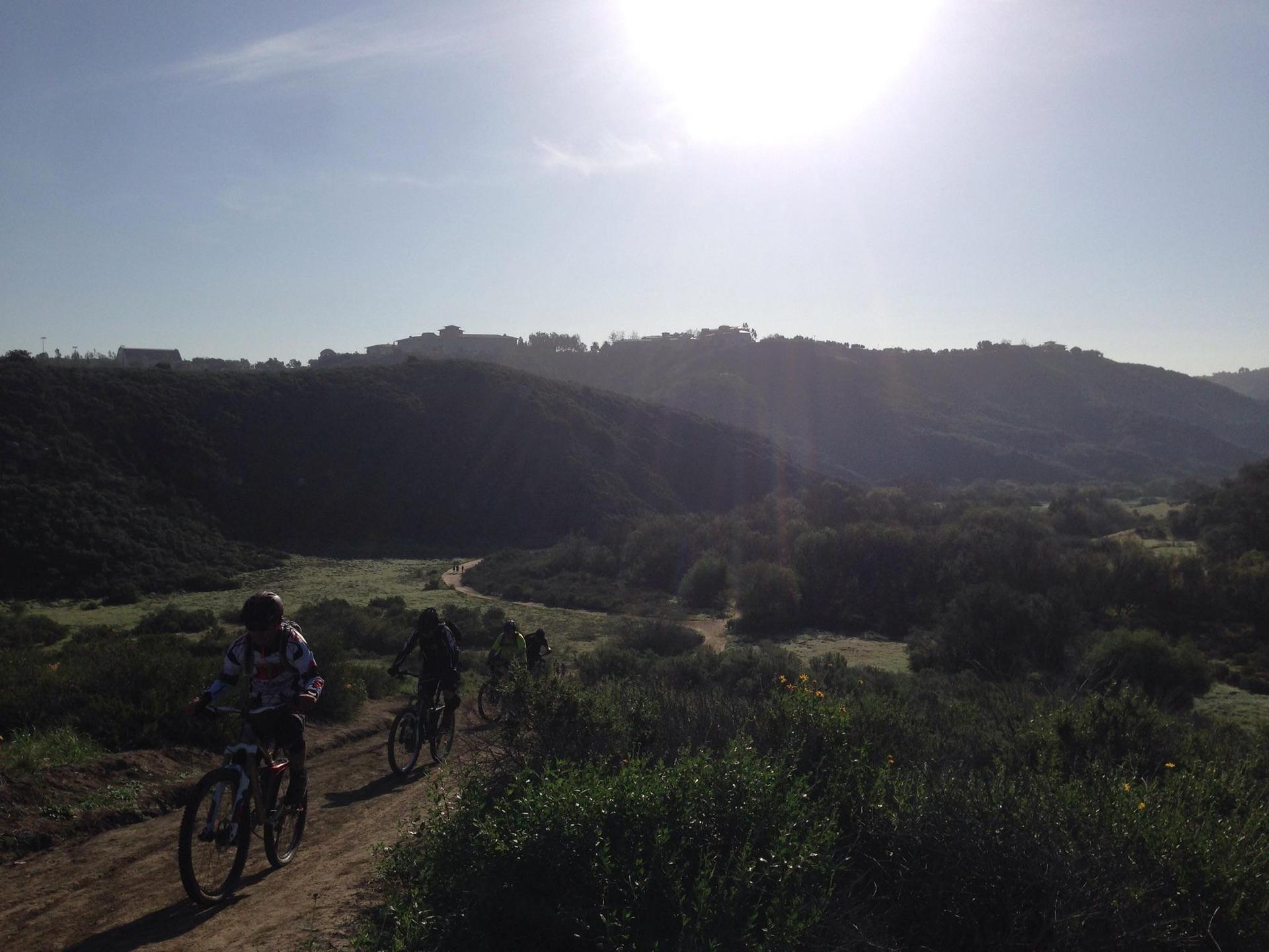 Mountain bikers on a dirt path with bright sun over hazy hills.