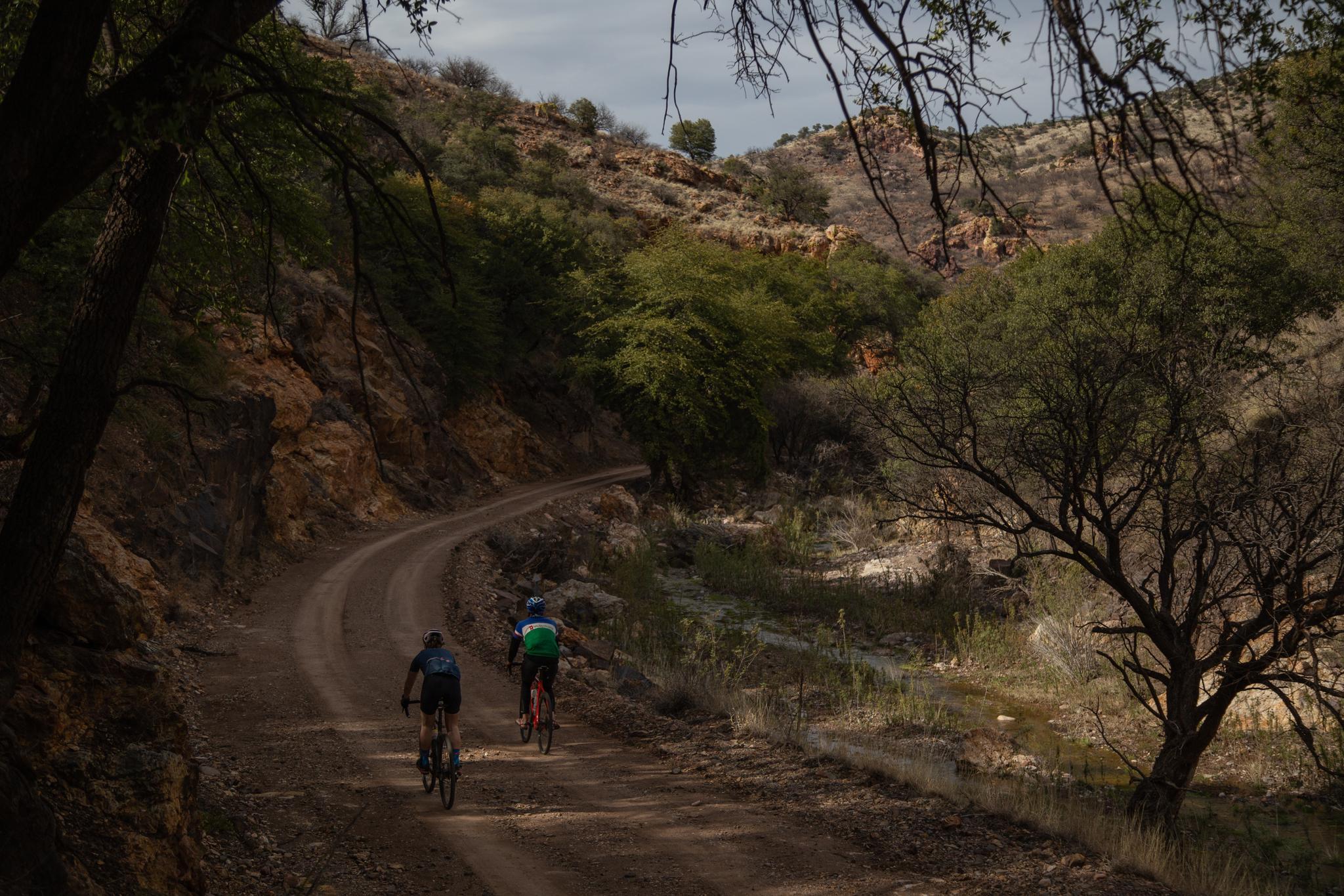 Two cyclists ride on a winding dirt road through a dry, hilly landscape.