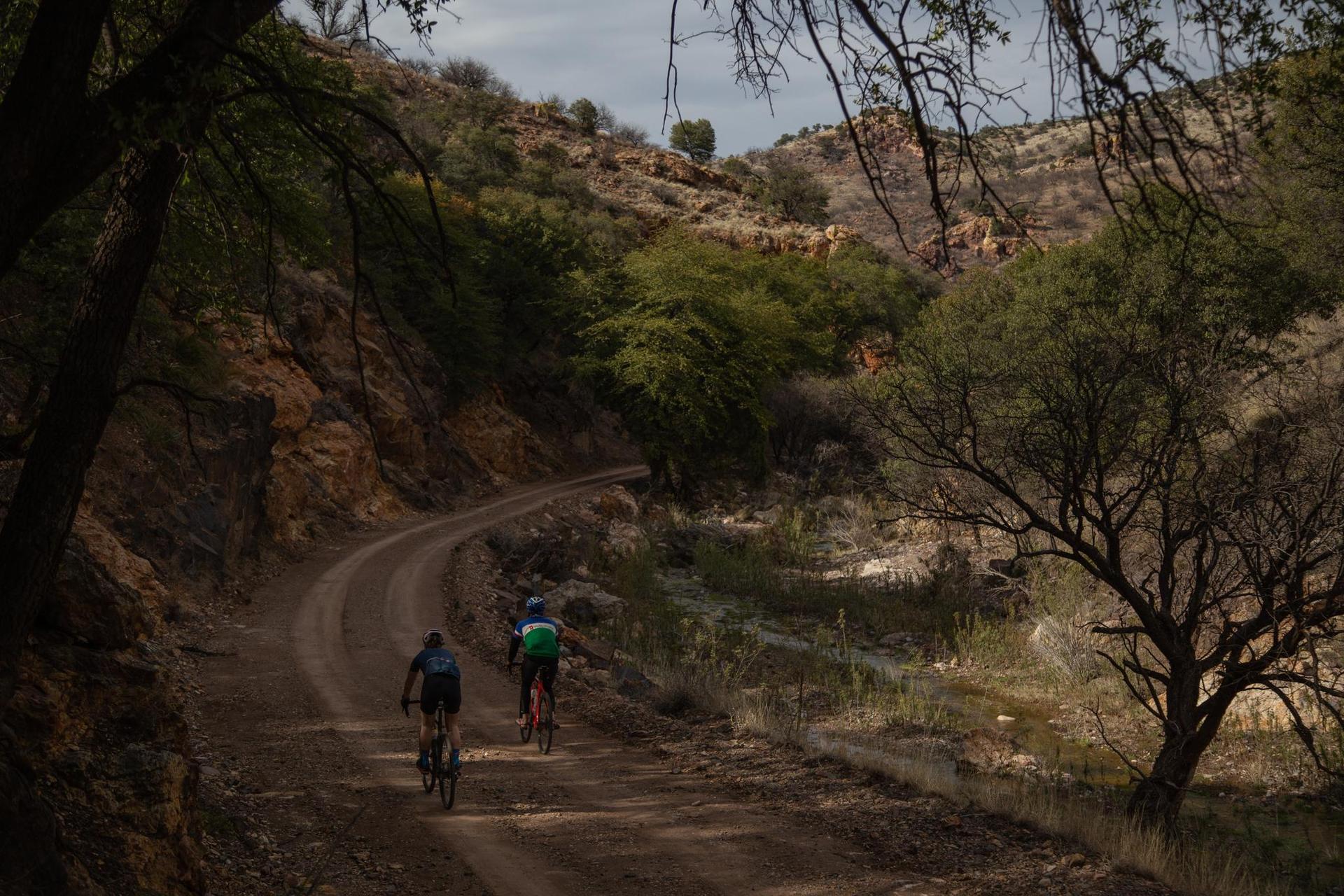 Two cyclists ride on a winding dirt road through a dry, hilly landscape.