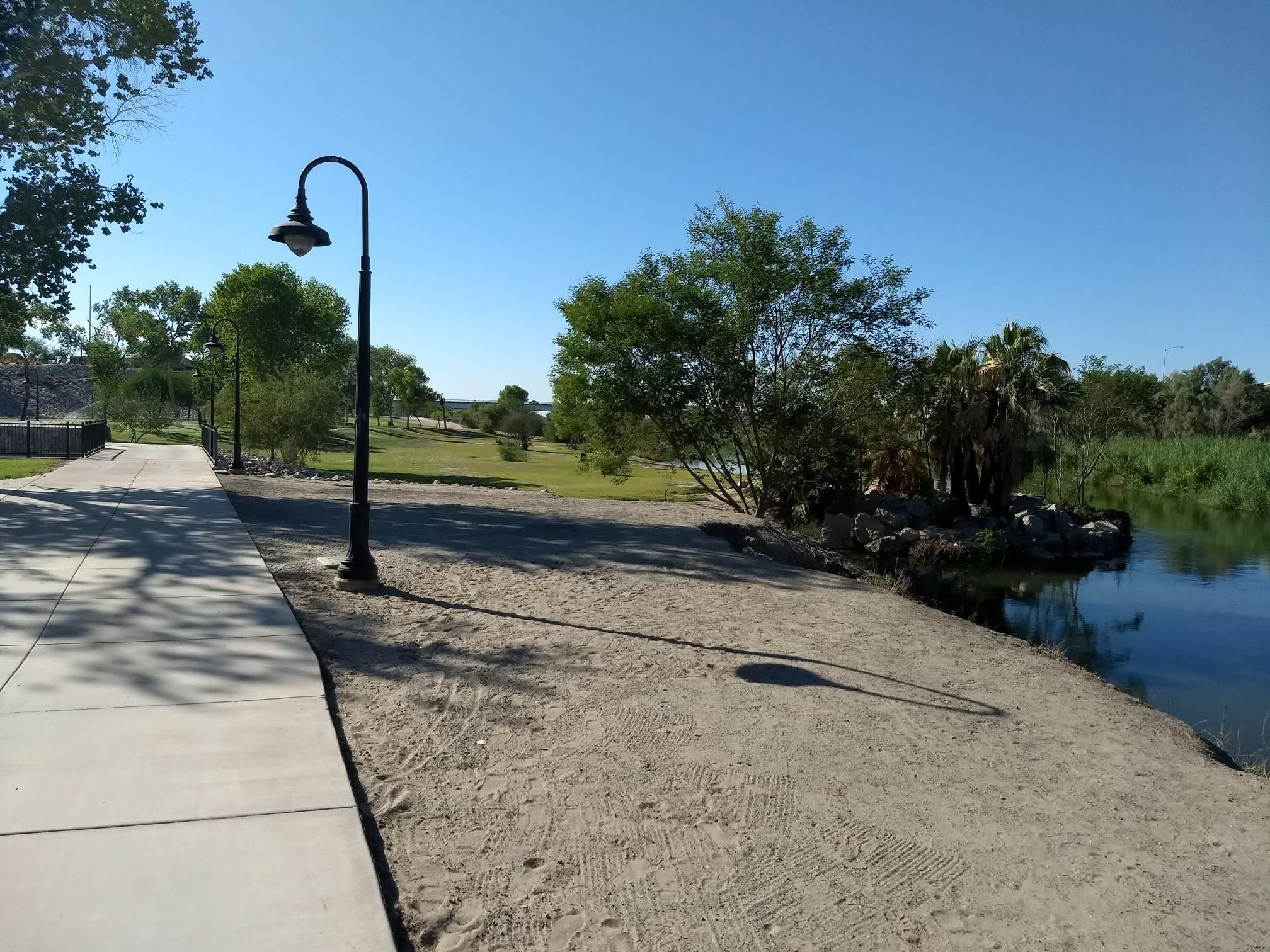 Paved path and sandy bank next to a calm river, with a lamppost. - Pivot Point Park – Riverside Park loop from Centennial Middle School