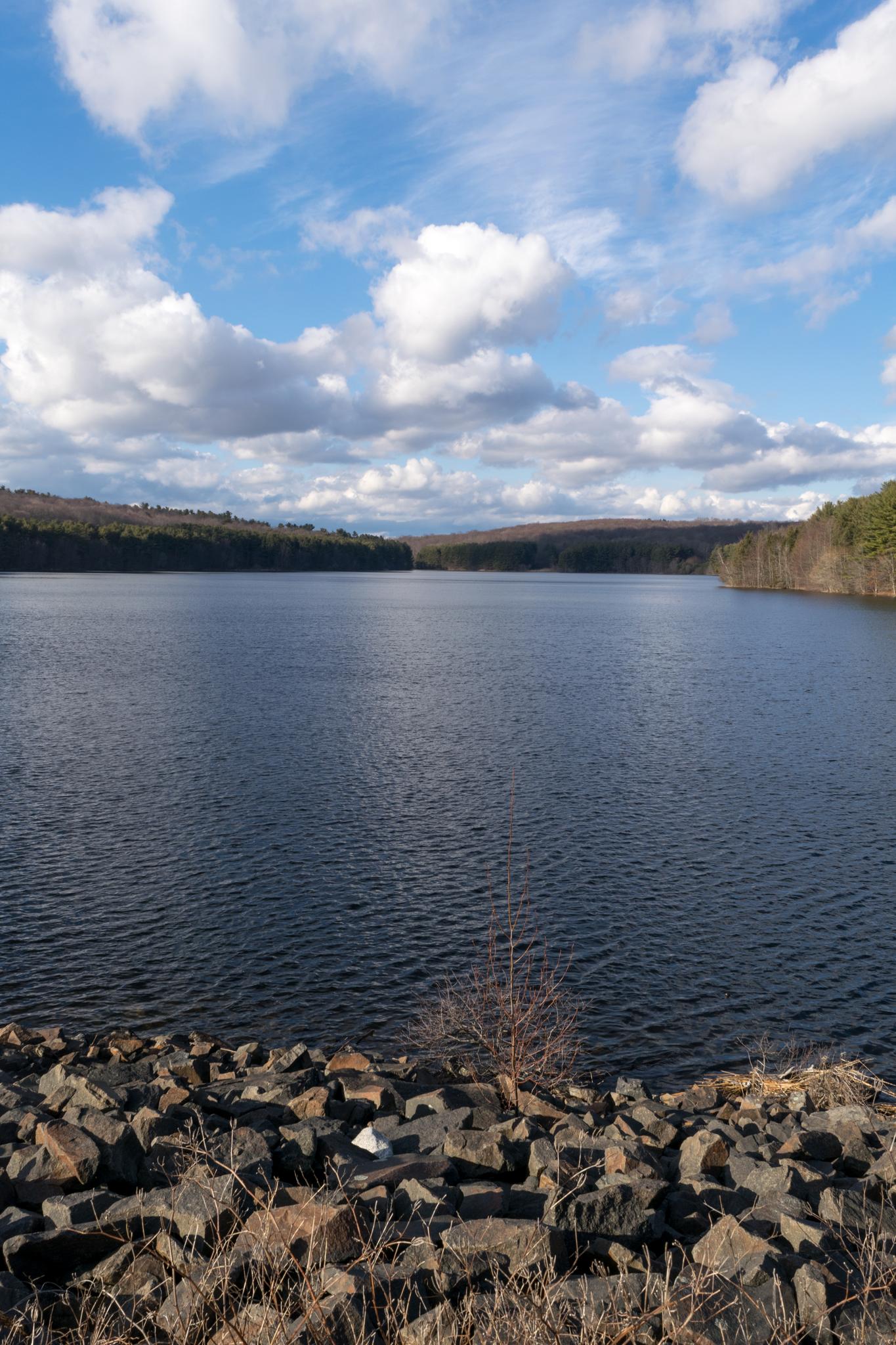 A wide lake with a rocky shore under a bright, partly cloudy sky.