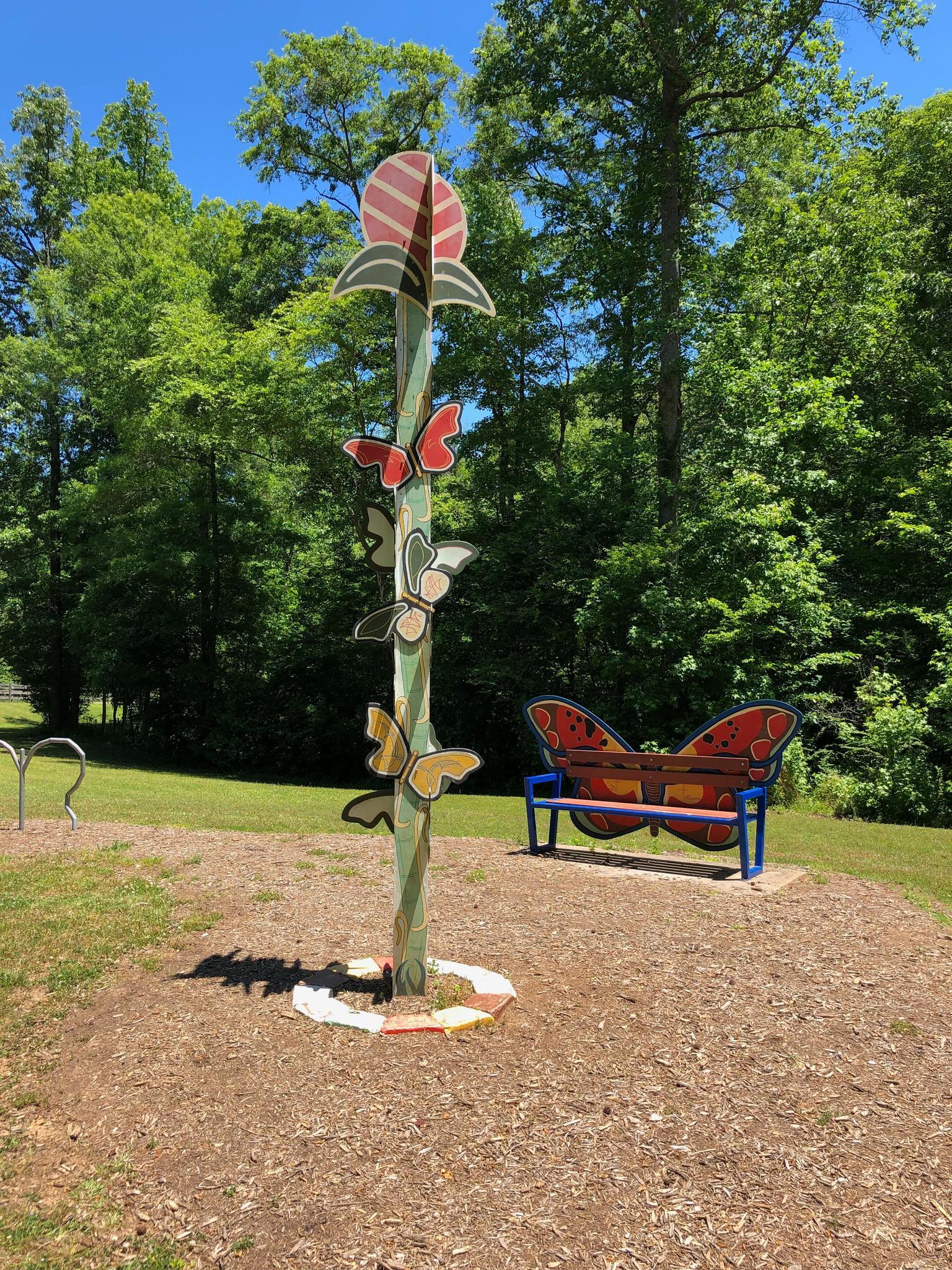 Colorful butterfly sculpture and bench in a sunny, wooded park setting.