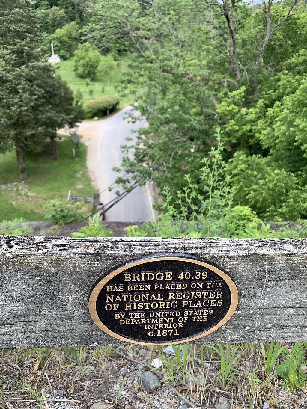 A bronze plaque on a wooden railing overlooks a road and green valley.