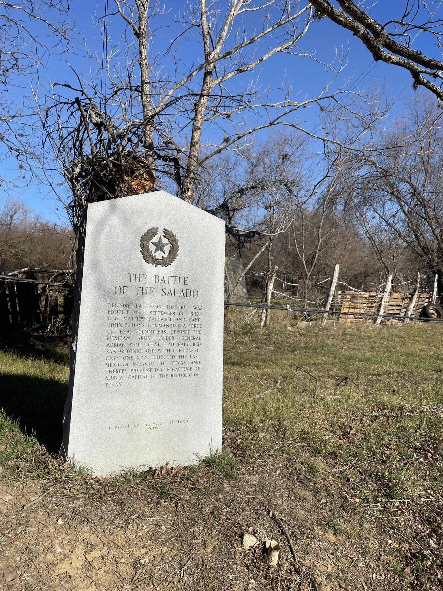 Stone historical marker in a sunny grassy field with bare trees and a fence.