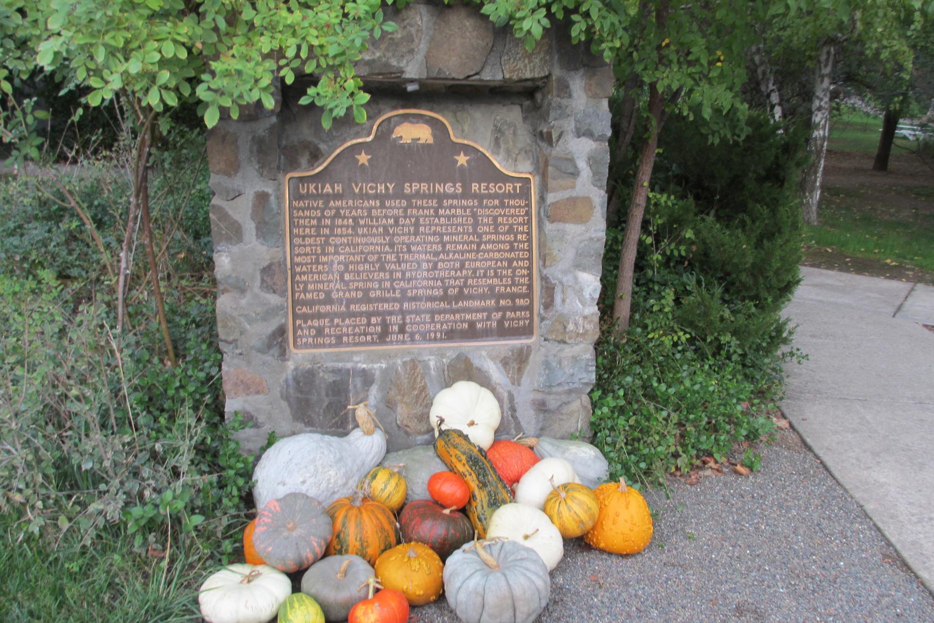 Stone monument with a plaque, surrounded by colorful gourds and pumpkins.