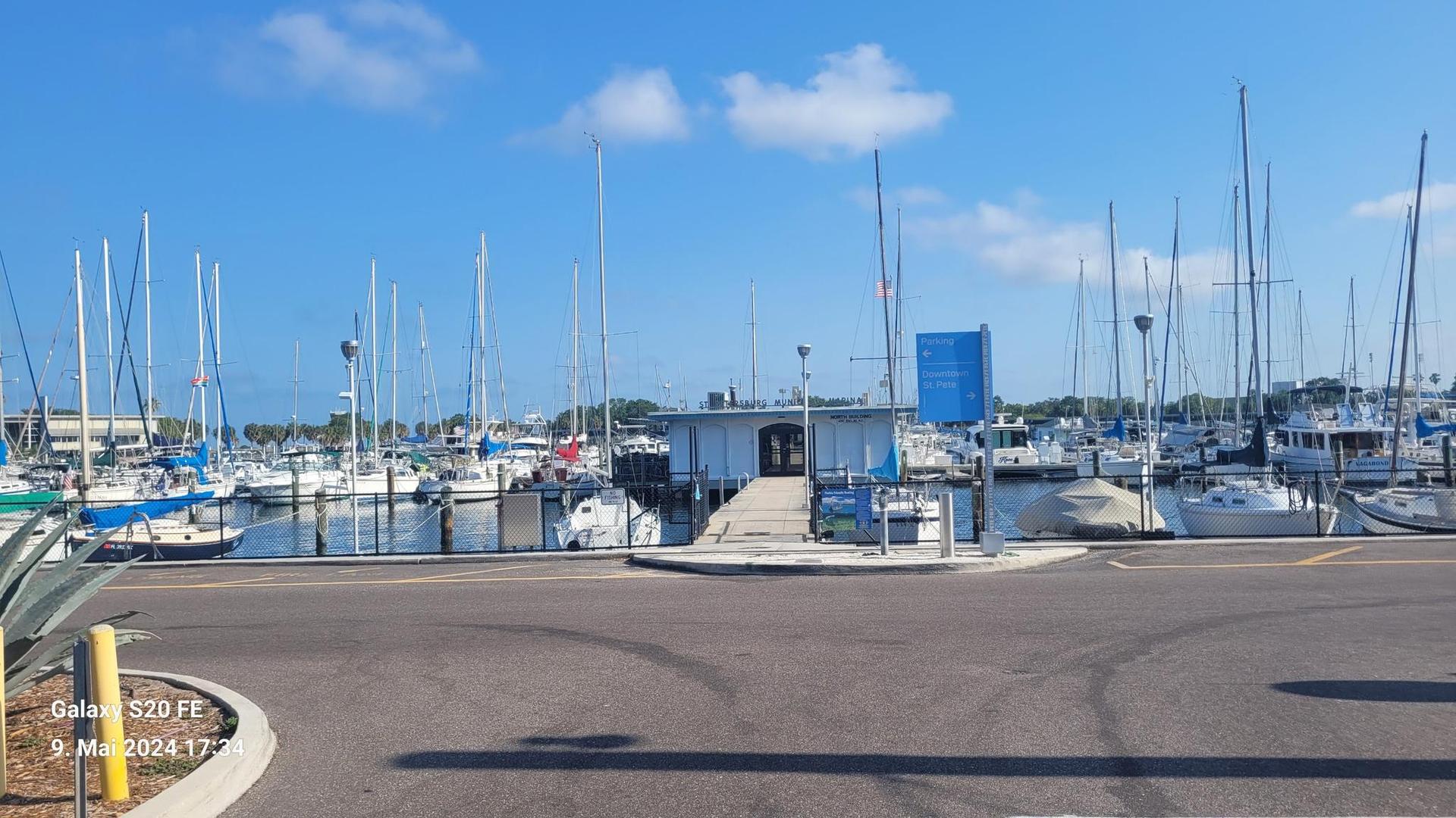 Sunny marina filled with sailboats, with a small building and parking lot.
