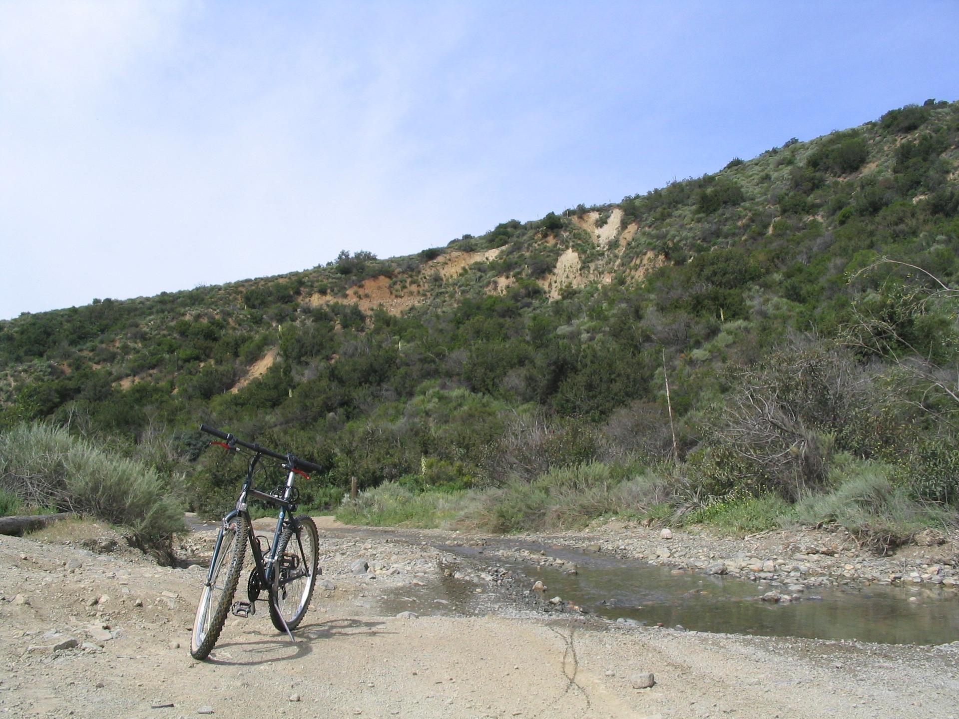 Bicycle on a muddy dirt road beside a green hillside under cloudy sky.