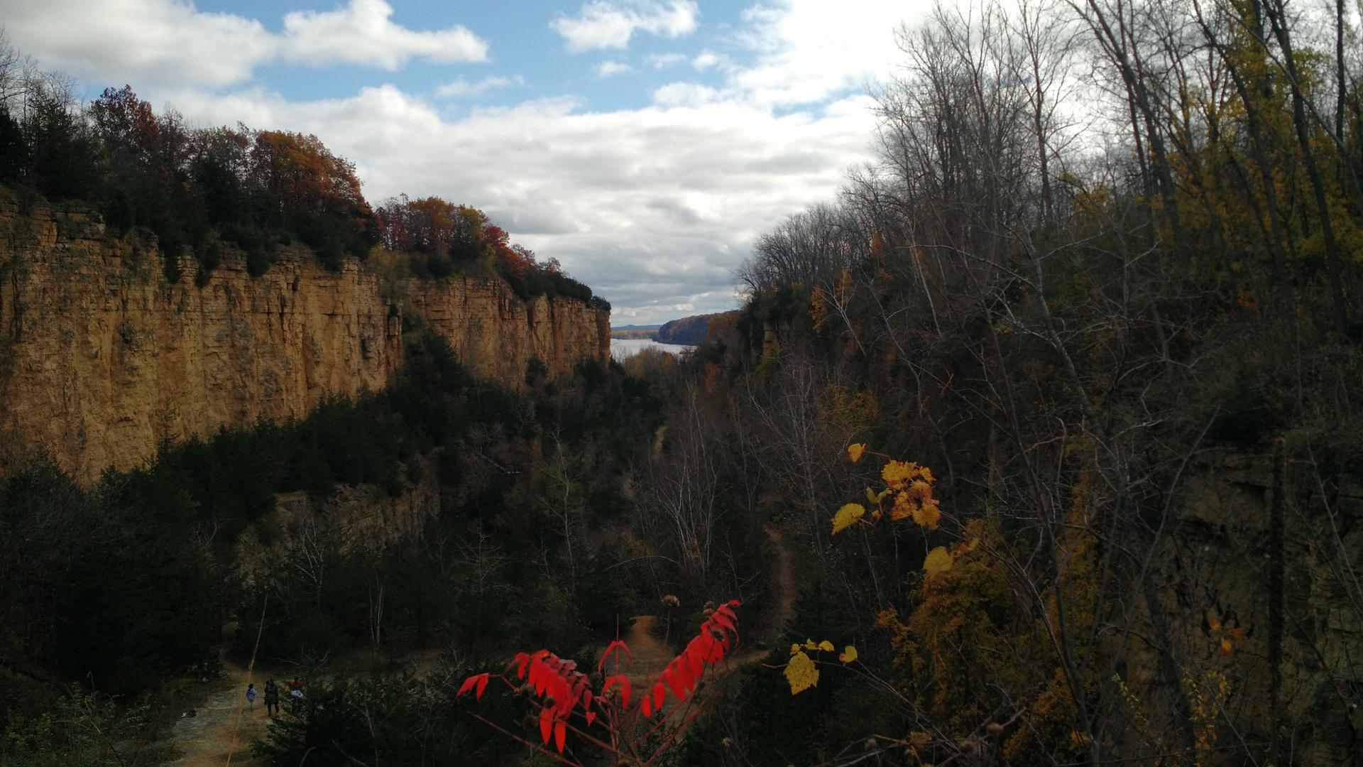 A scenic canyon with rocky cliffs, autumn trees, and distant water under a cloudy sky.
