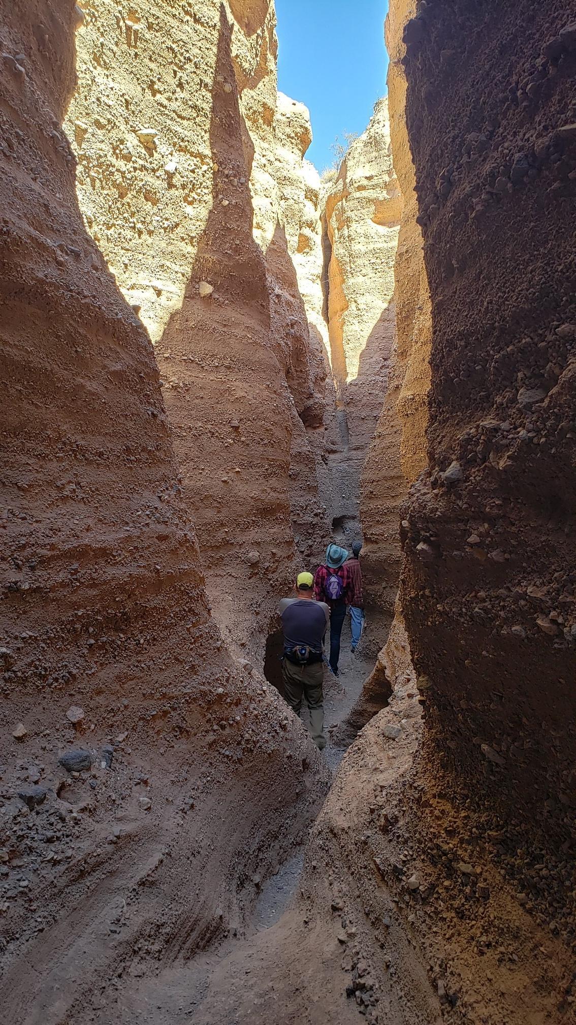 Hikers navigating a narrow, sunlit slot canyon with tall, rocky walls.