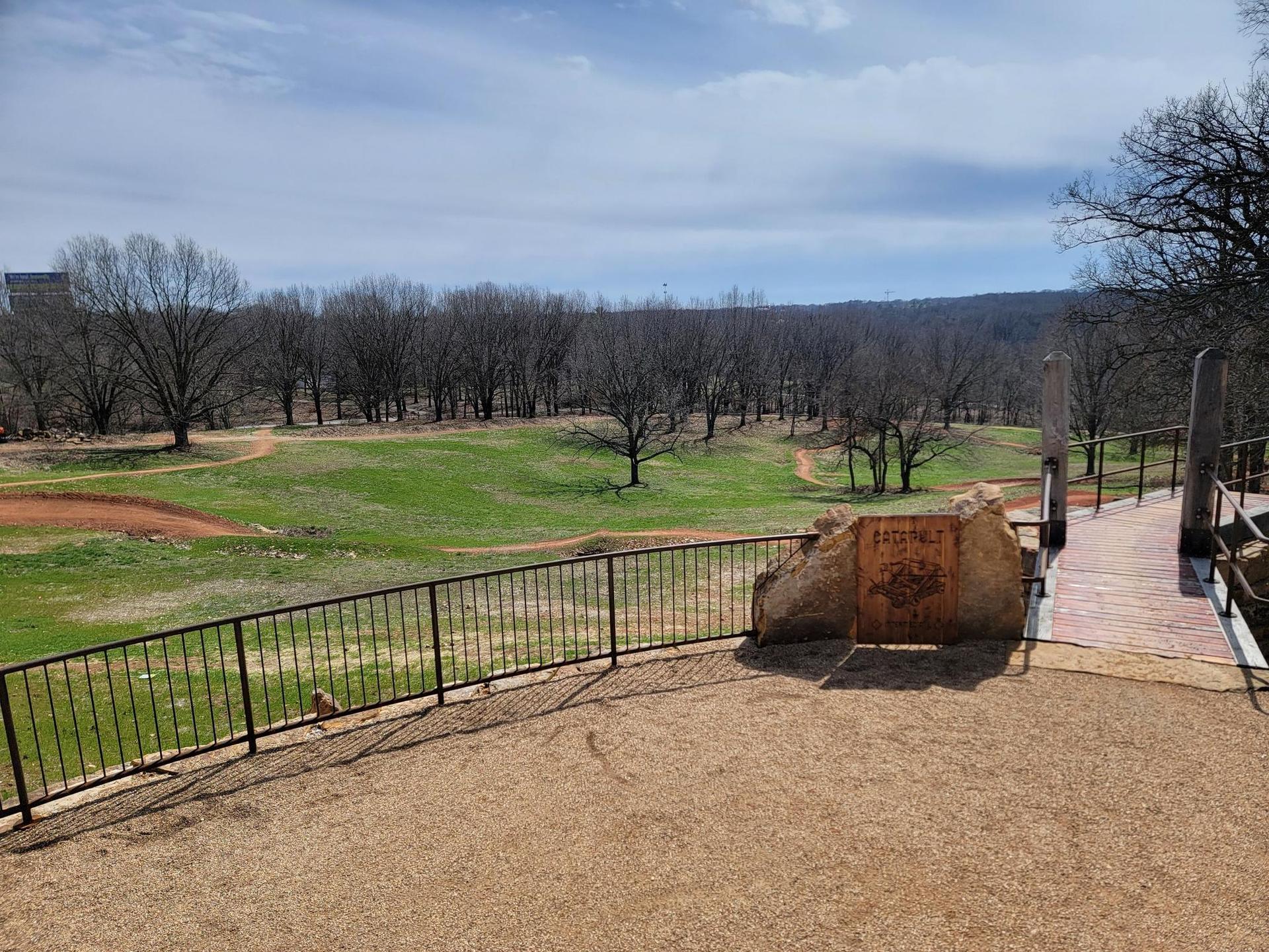 Overlook with fence and bridge, viewing a green field and bare trees.
