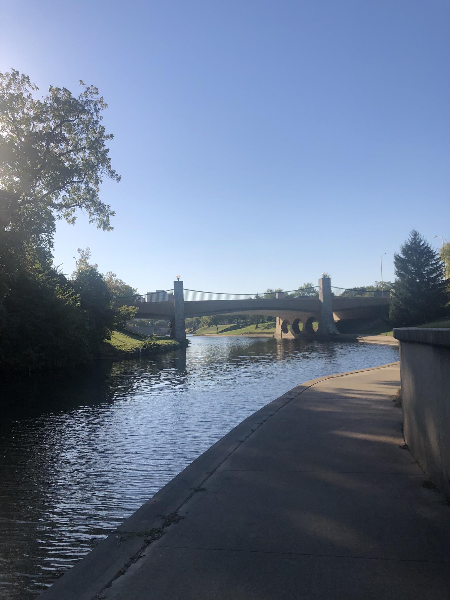 A sunny river with a concrete path and a large bridge in the distance.