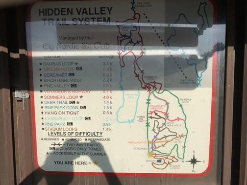 A sunny outdoor trail map display board with a person's shadow.