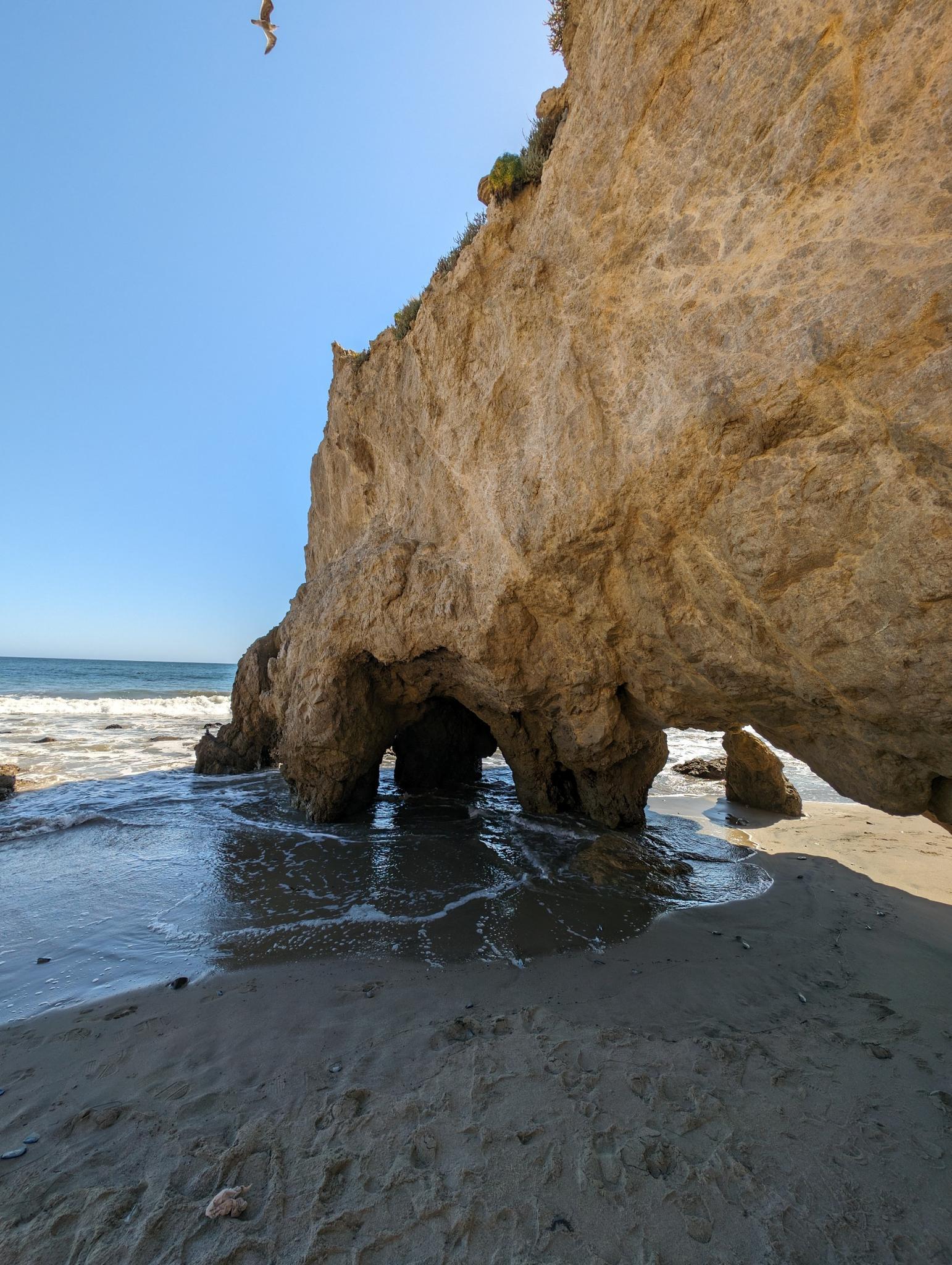 Ocean water flowing into a large rock cave on a sunny sandy beach.