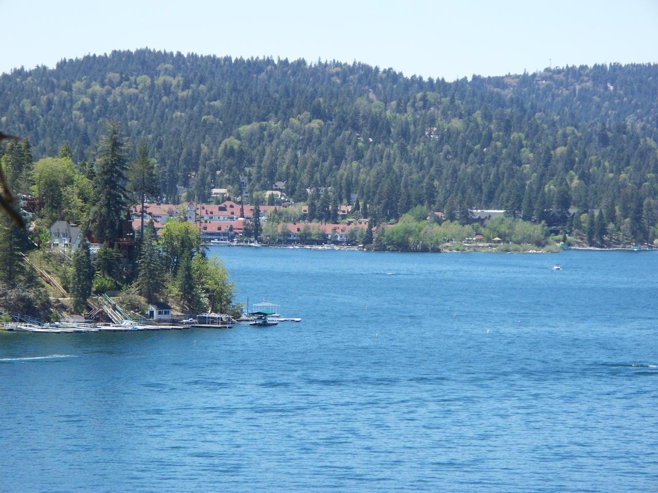 Expansive blue lake with green forested shores and distant houses on a clear day.