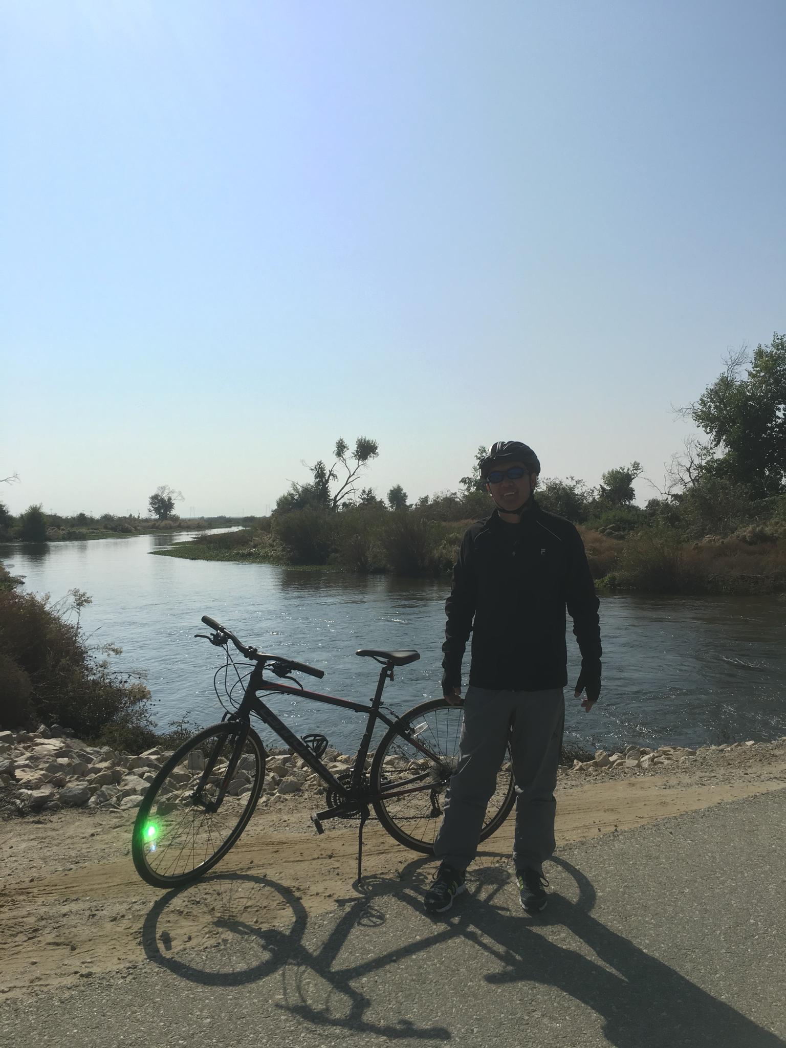 Person stands with bicycle by a sunny river with trees.