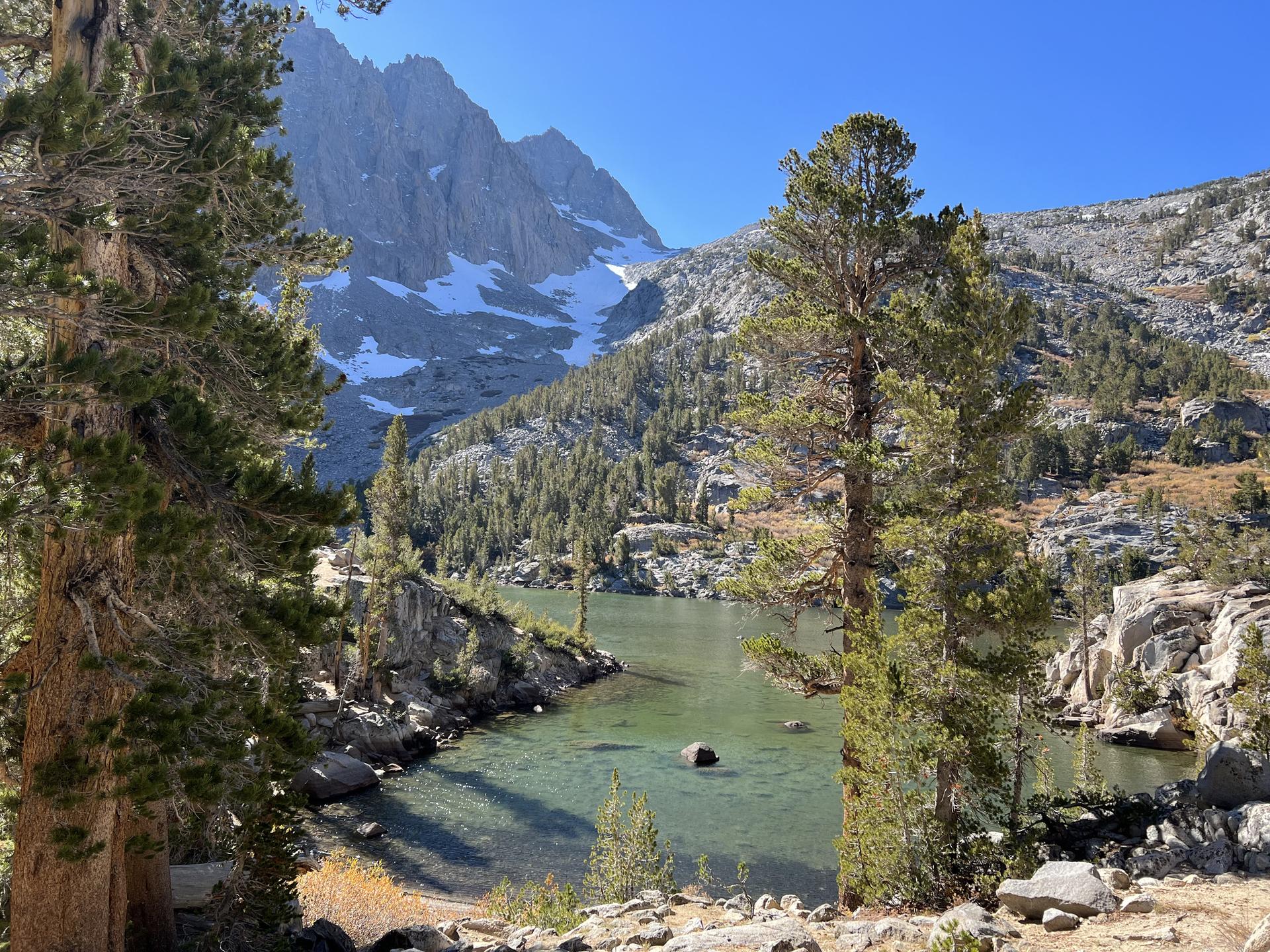Clear mountain lake with pine trees and snow-dusted peaks under bright sun.