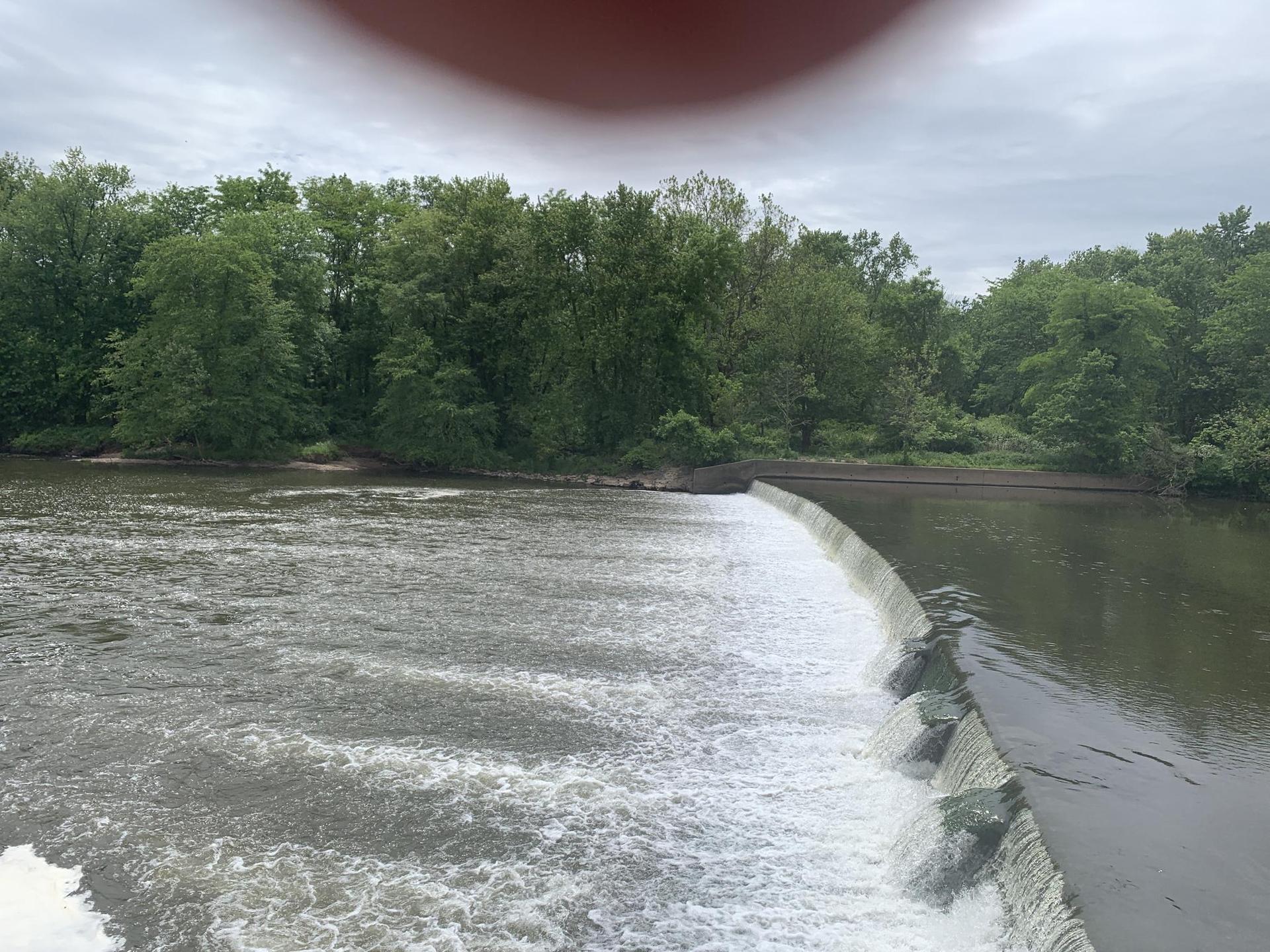 Overcast view of a river flowing over a low dam, surrounded by green trees.