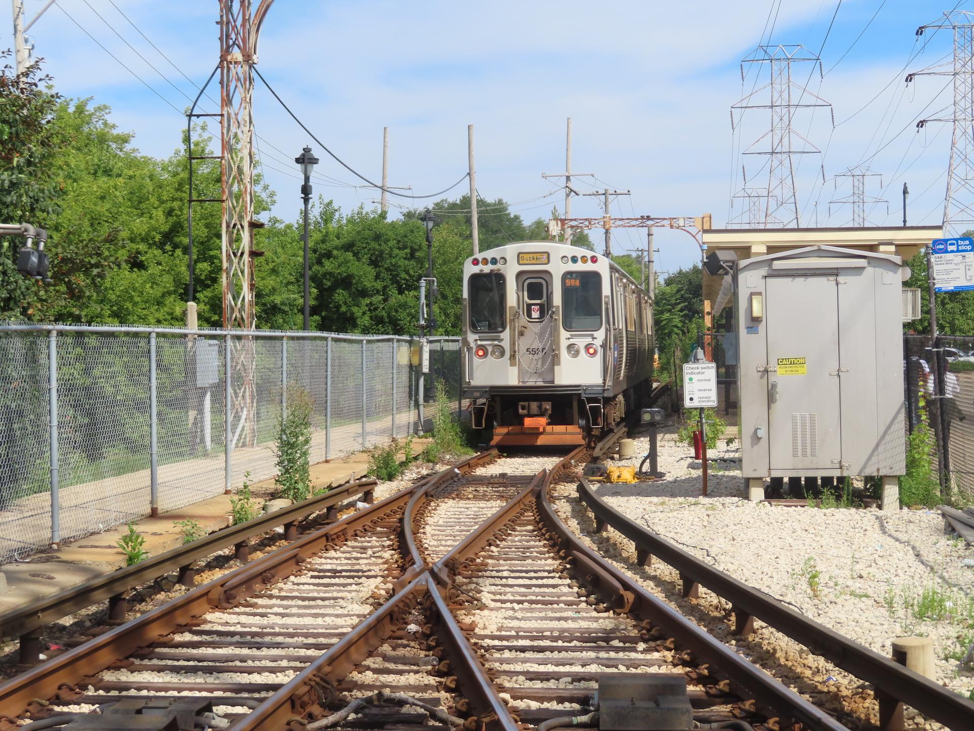 Train on tracks with power lines, a fence, and green trees on a sunny day.