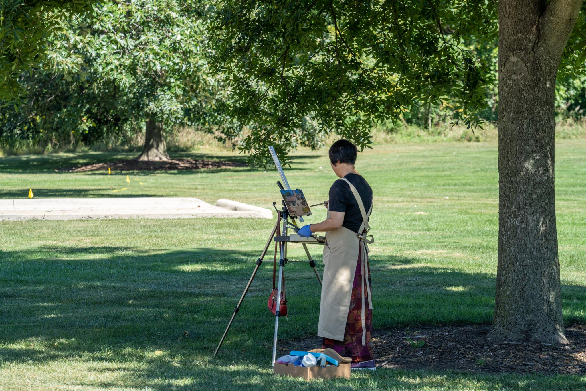 Person painting outdoors at an easel under a tree on a sunny day.