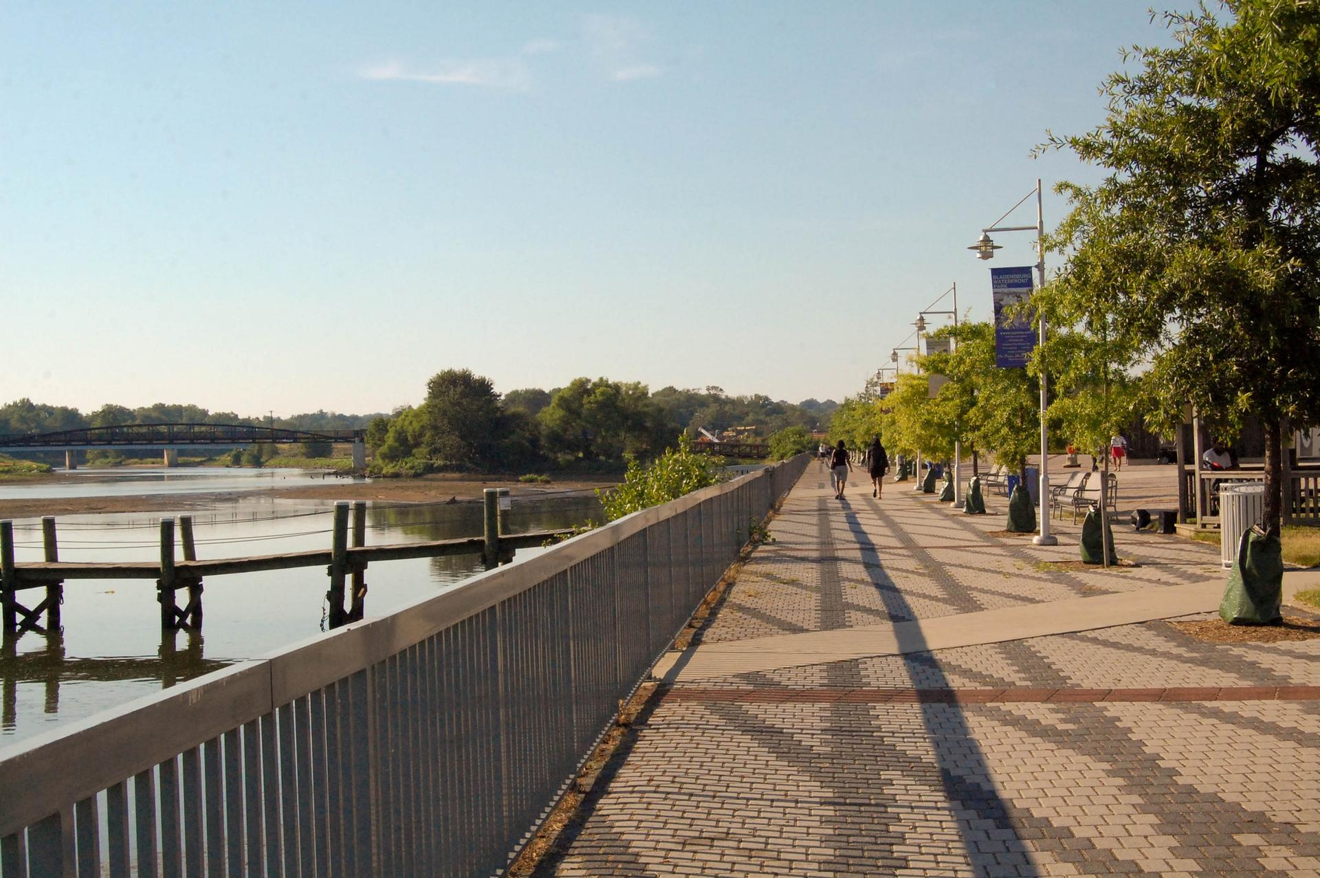 People walking on a sunny boardwalk next to a river with docks and a distant bridge.