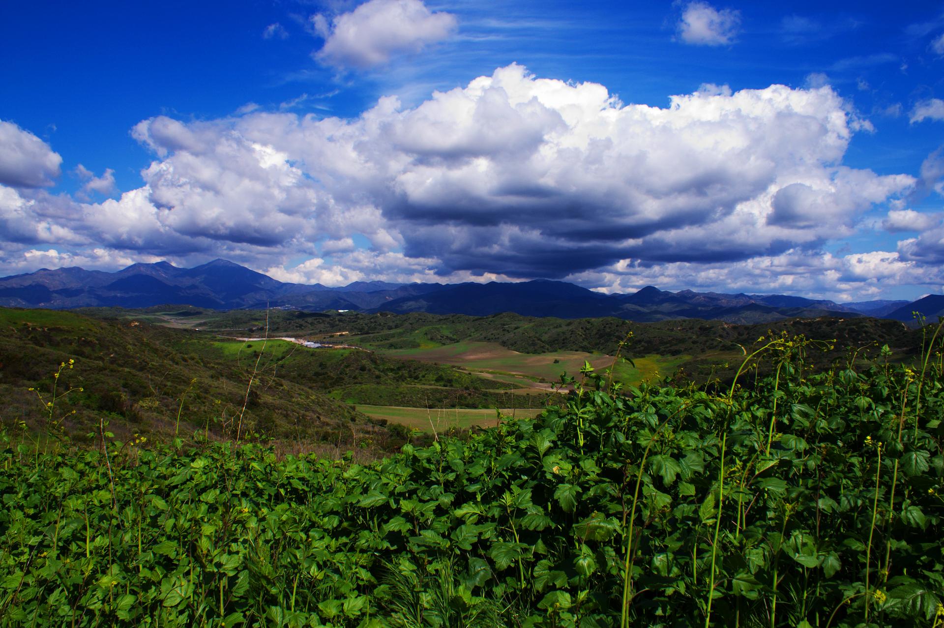 Expansive green landscape with rolling hills and mountains under a dynamic cloudy sky.