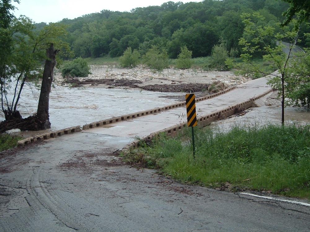 Flooded concrete bridge partially submerged by muddy, fast-flowing river water.