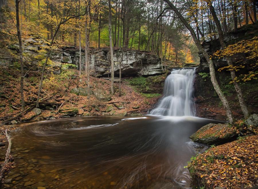 Waterfall cascades into a dark pool surrounded by colorful autumn trees and rocks.