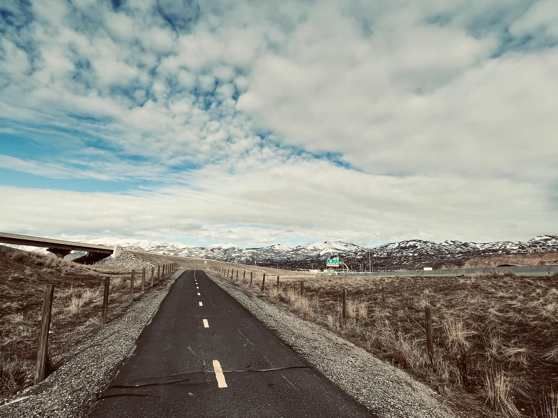 Paved road through dry landscape towards snow-dusted mountains under a cloudy sky.