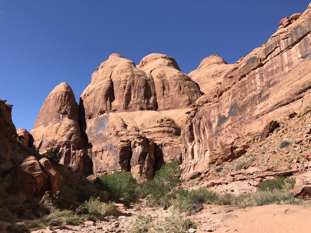 Towering red rock formations and cliffs under a clear blue sky, with green shrubs.