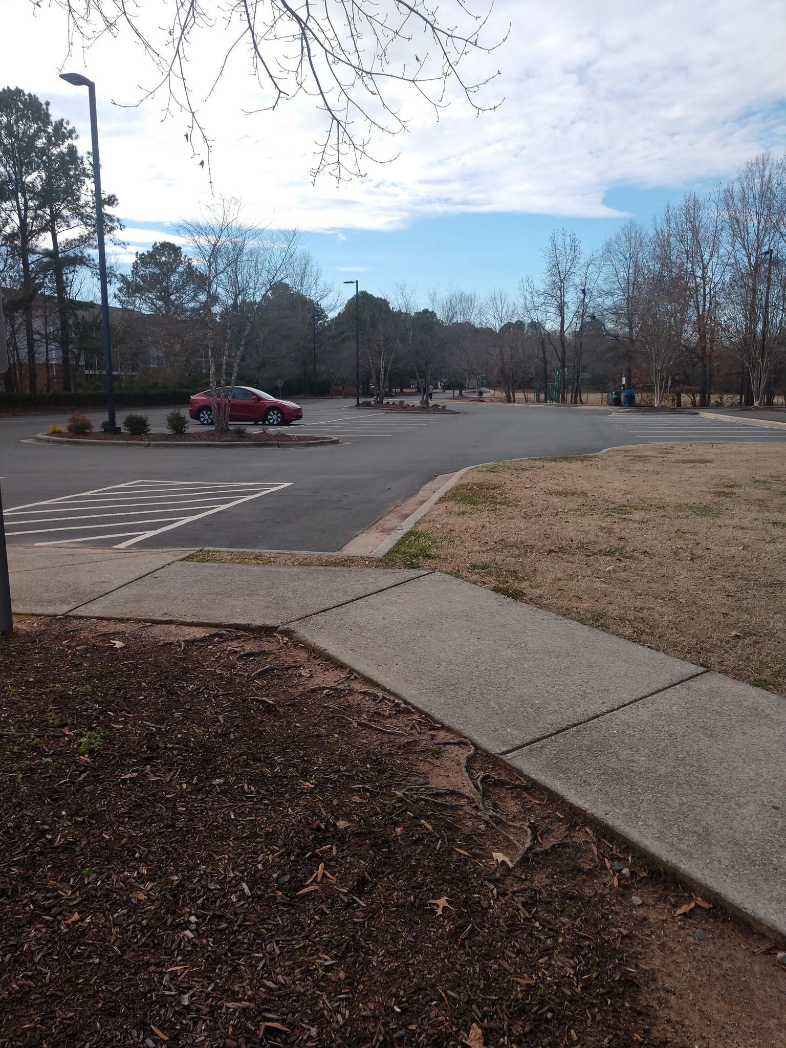 A parking lot with a red car, bare trees, and a sidewalk under a bright, cloudy sky.