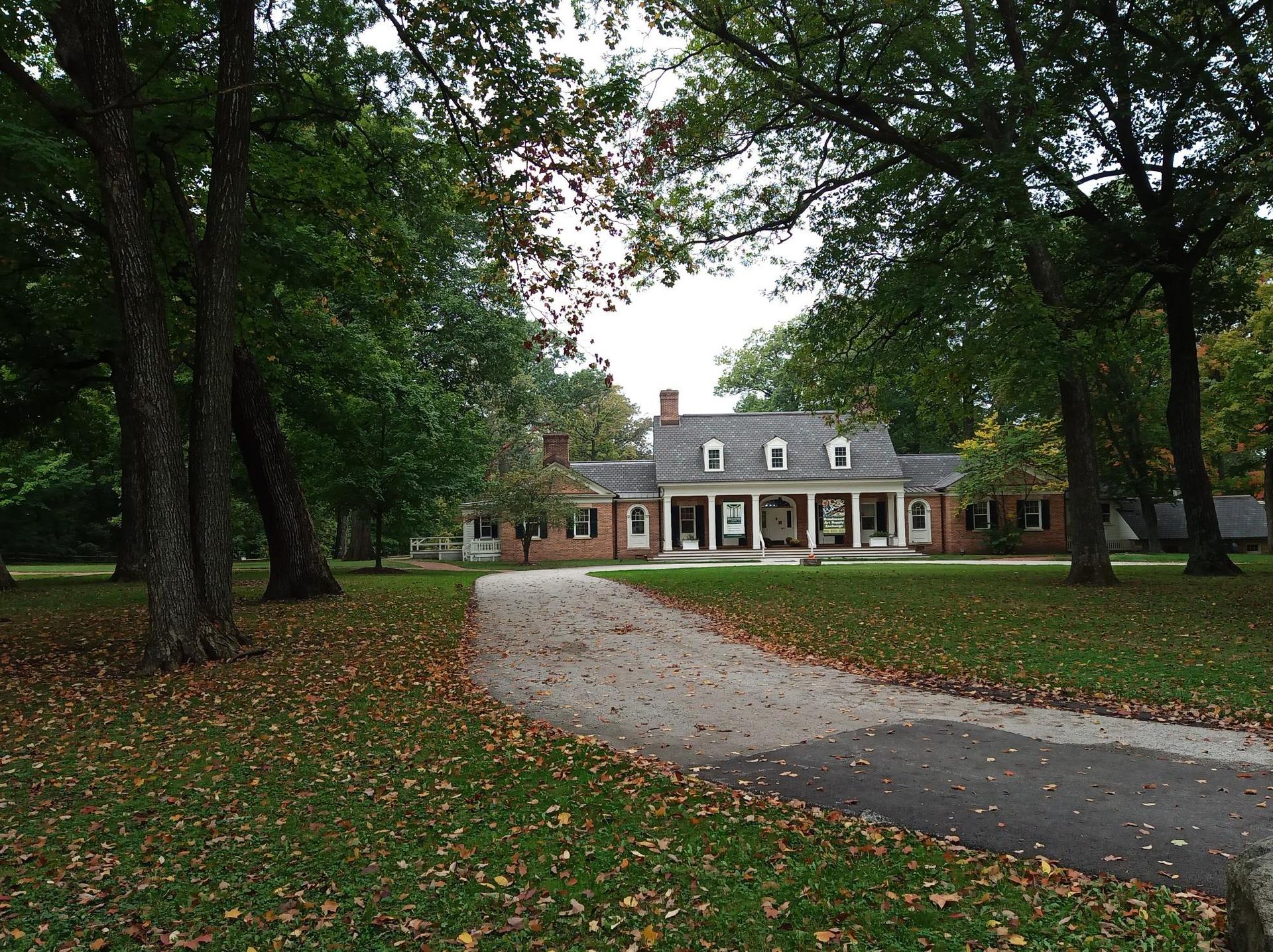 Long brick house with white columns at end of a driveway, surrounded by trees.