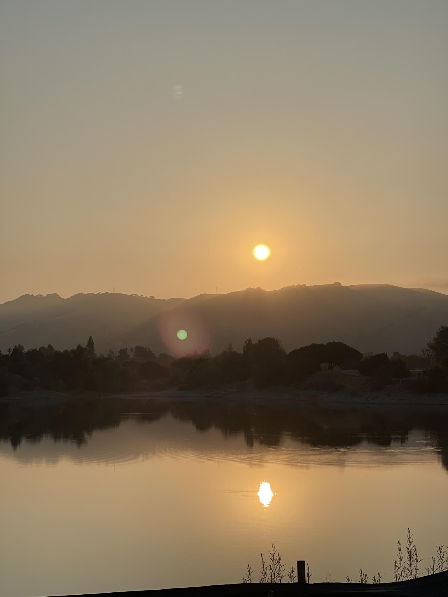 Golden sunset over a calm lake reflecting the sun, with silhouetted hills.