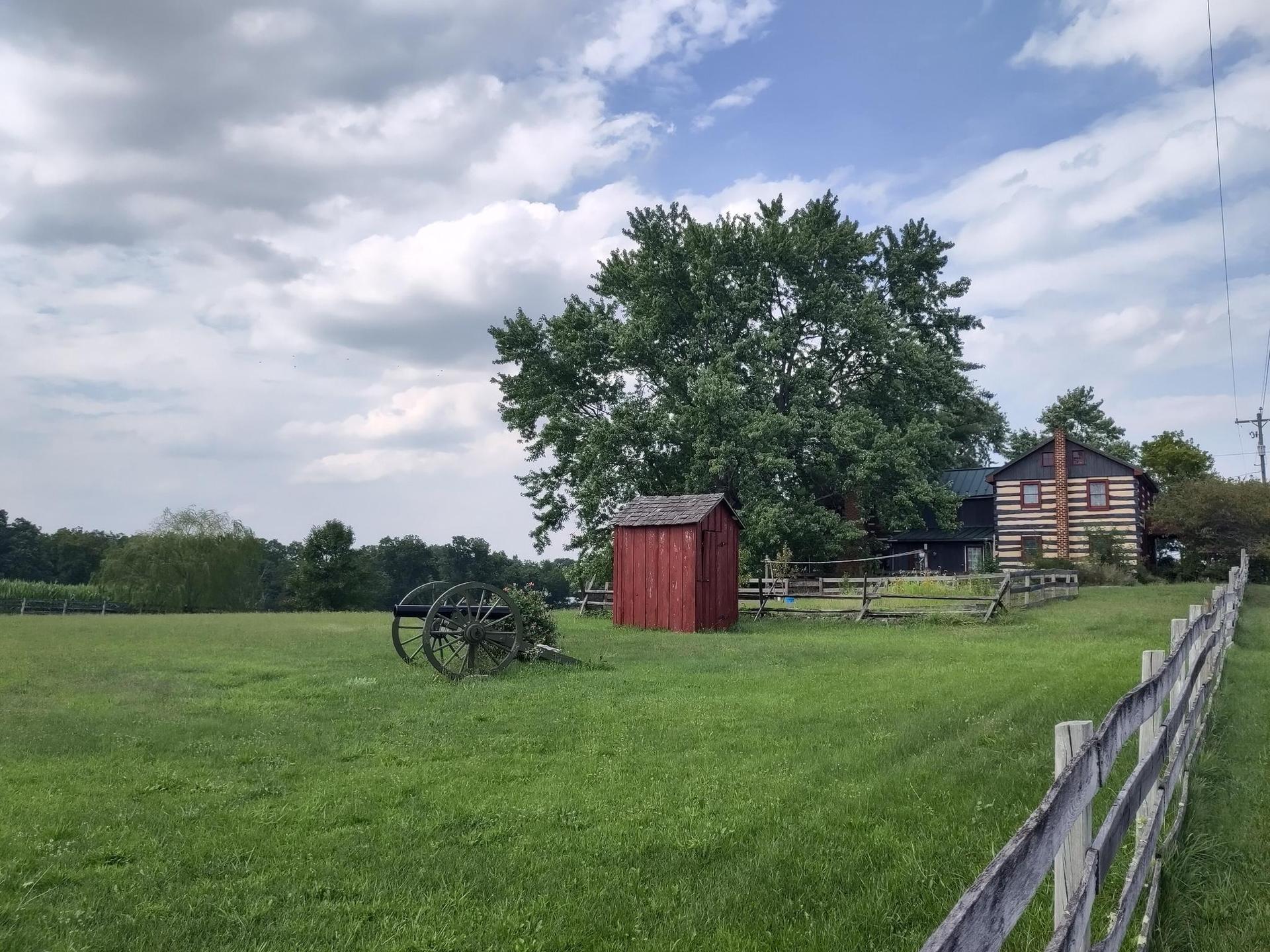 A red shed, log cabin, and wagon in a grassy field under a cloudy sky.