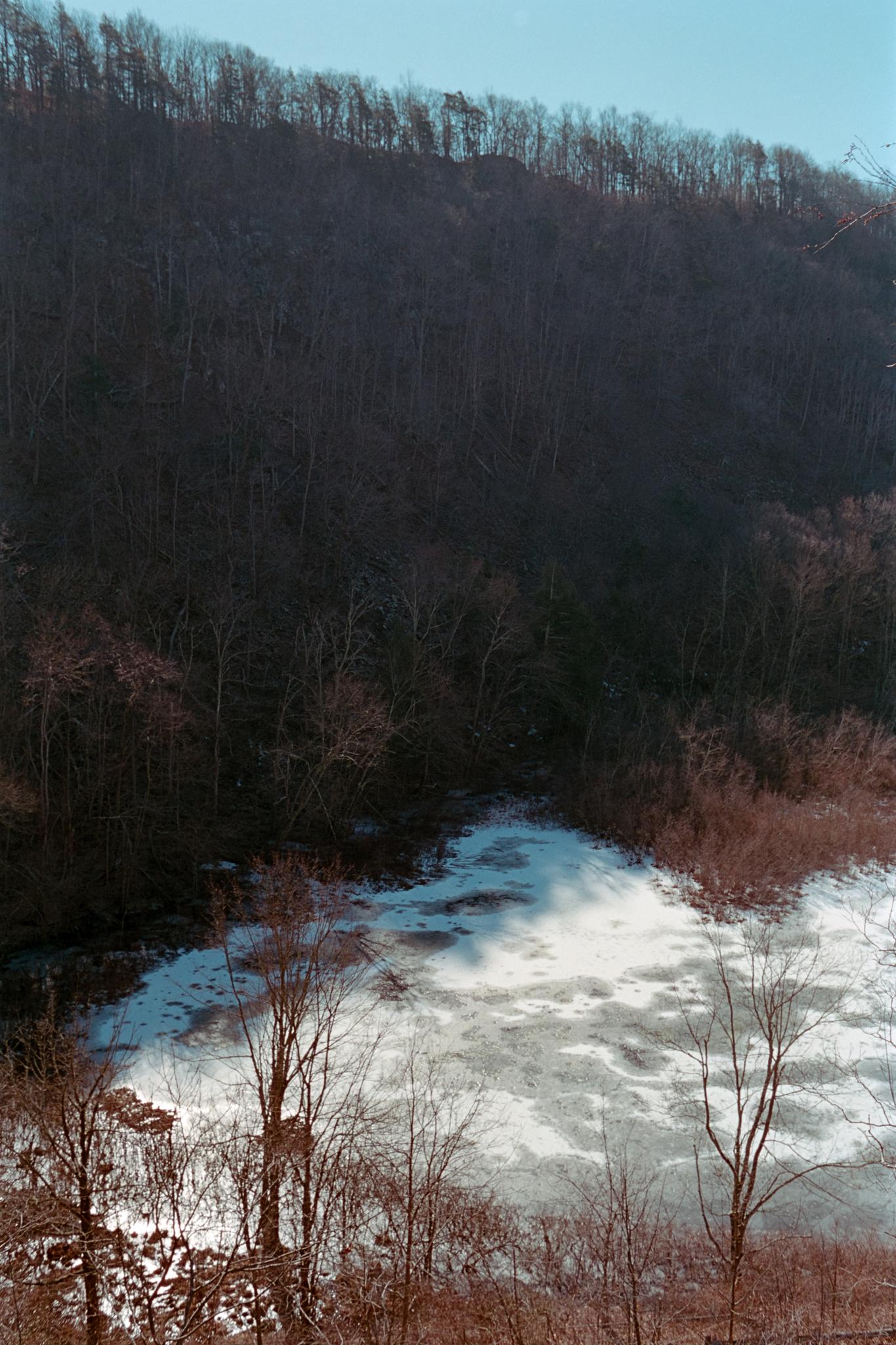 Dark wooded hillside overlooking a partially frozen body of water in winter.