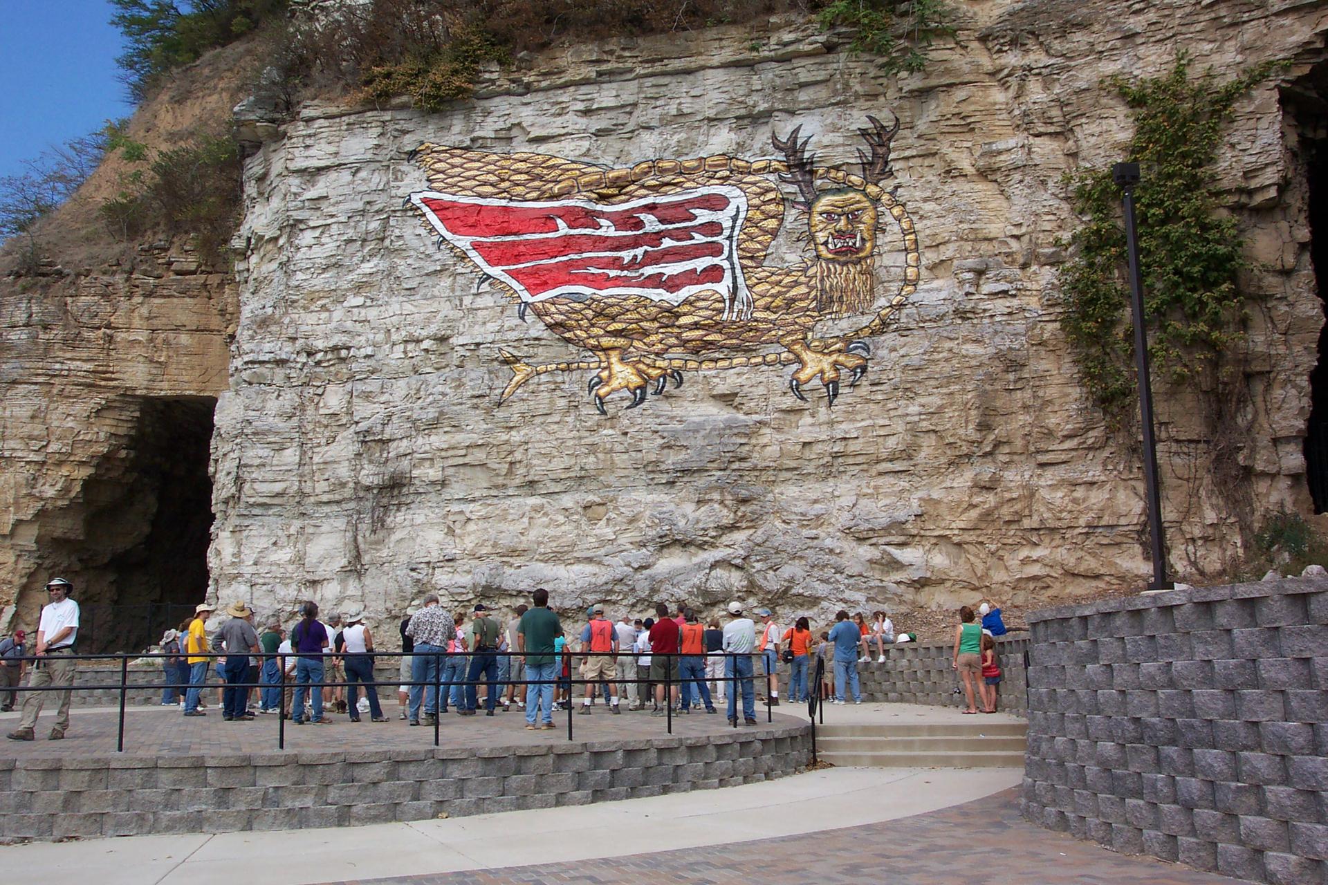 People view a large mythical creature painted on a sunny rock cliff face.