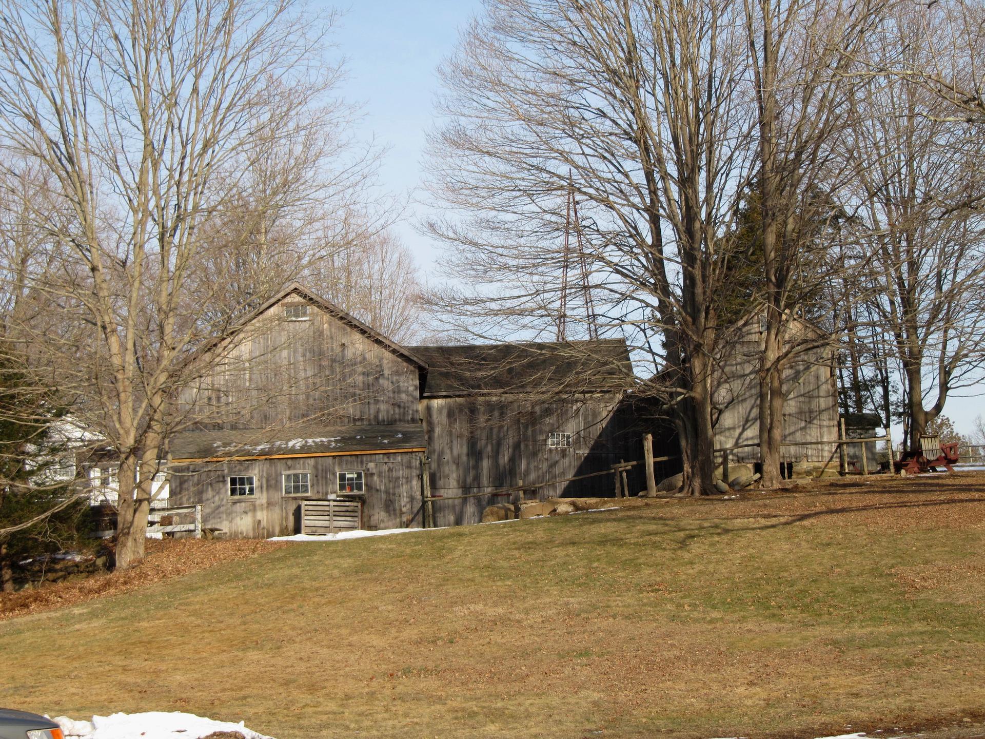 Weathered barn and silo among bare trees on a sunny, grassy slope with snow patches.